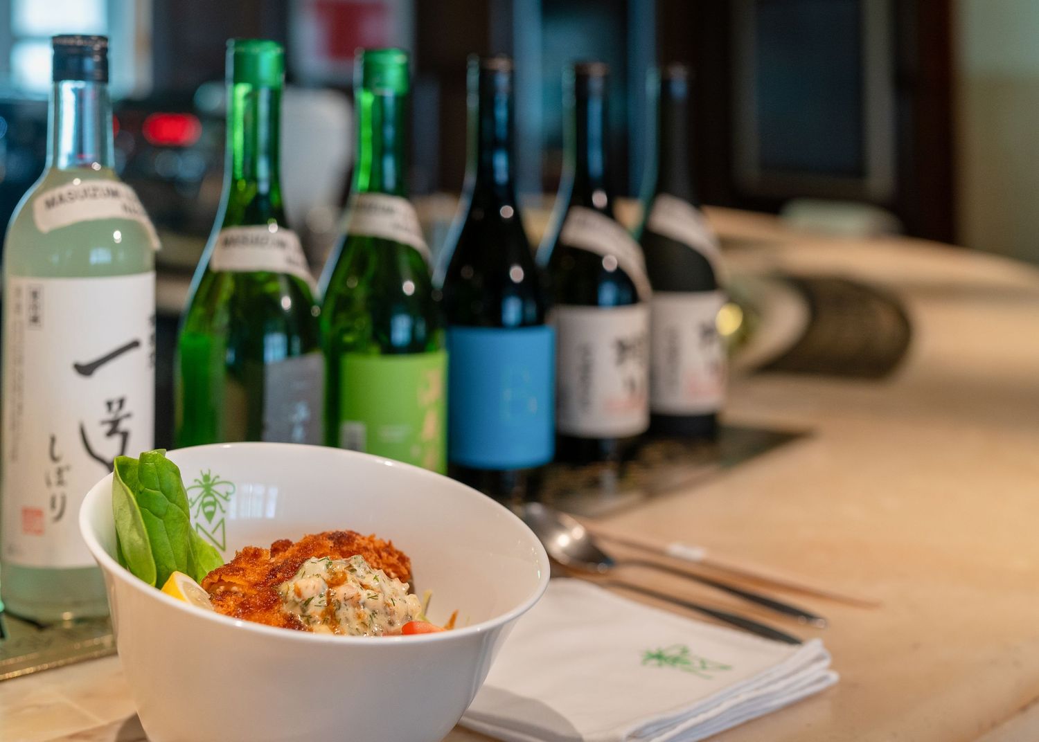 A bowl of rice dish with fresh basil sits on a counter beside a row of Japanese sake and wine bottles.