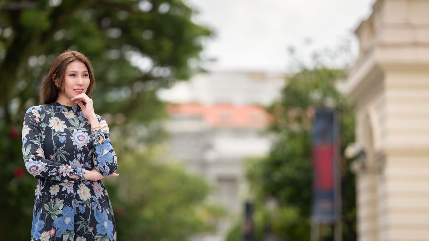 A stylish Asian woman in a floral blue dress poses thoughtfully on a tree-lined urban street.