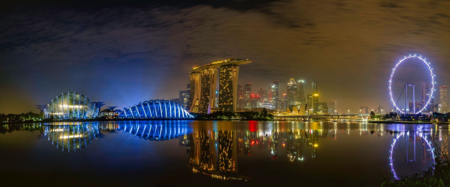 Panoramic Singapore Marina Bay skyline at night with glowing hotels, Ferris wheel, and vibrant reflections on calm water.