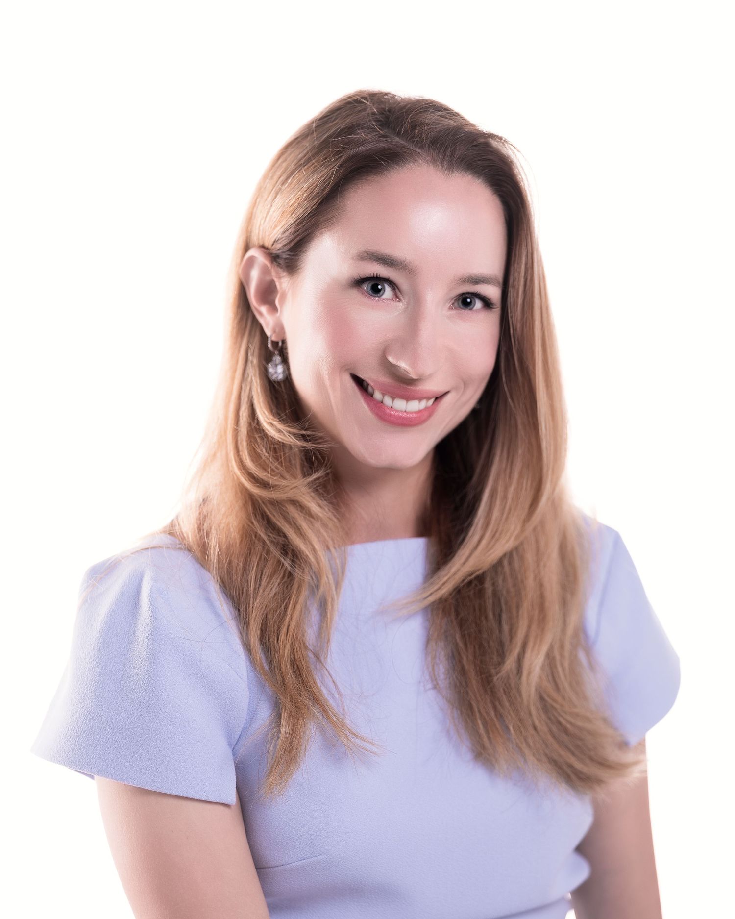 Smiling woman with long blonde hair wearing a light blue top and pearl earrings against a white background.