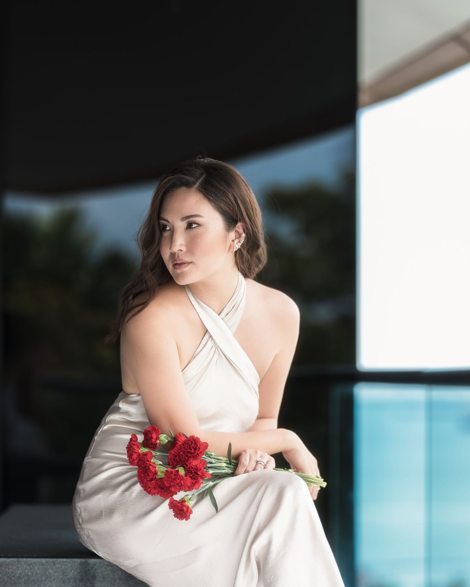 Elegant woman in white halter dress holding red carnations, seated outdoors near a pool in a sophisticated setting.
