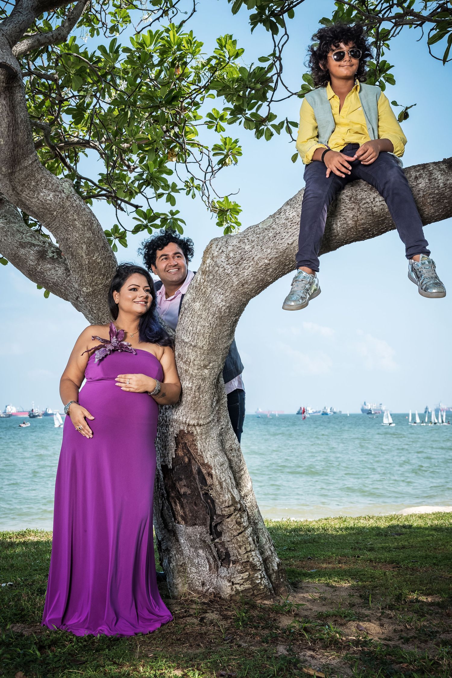 Pregnant woman in purple dress poses with family by a tree near the ocean, child sitting on branch above.