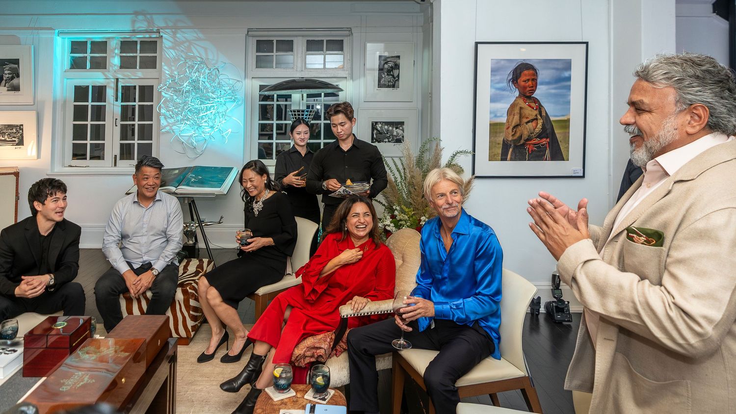 A man in a beige blazer addresses a seated group in a stylish room with framed artwork during a social gathering.