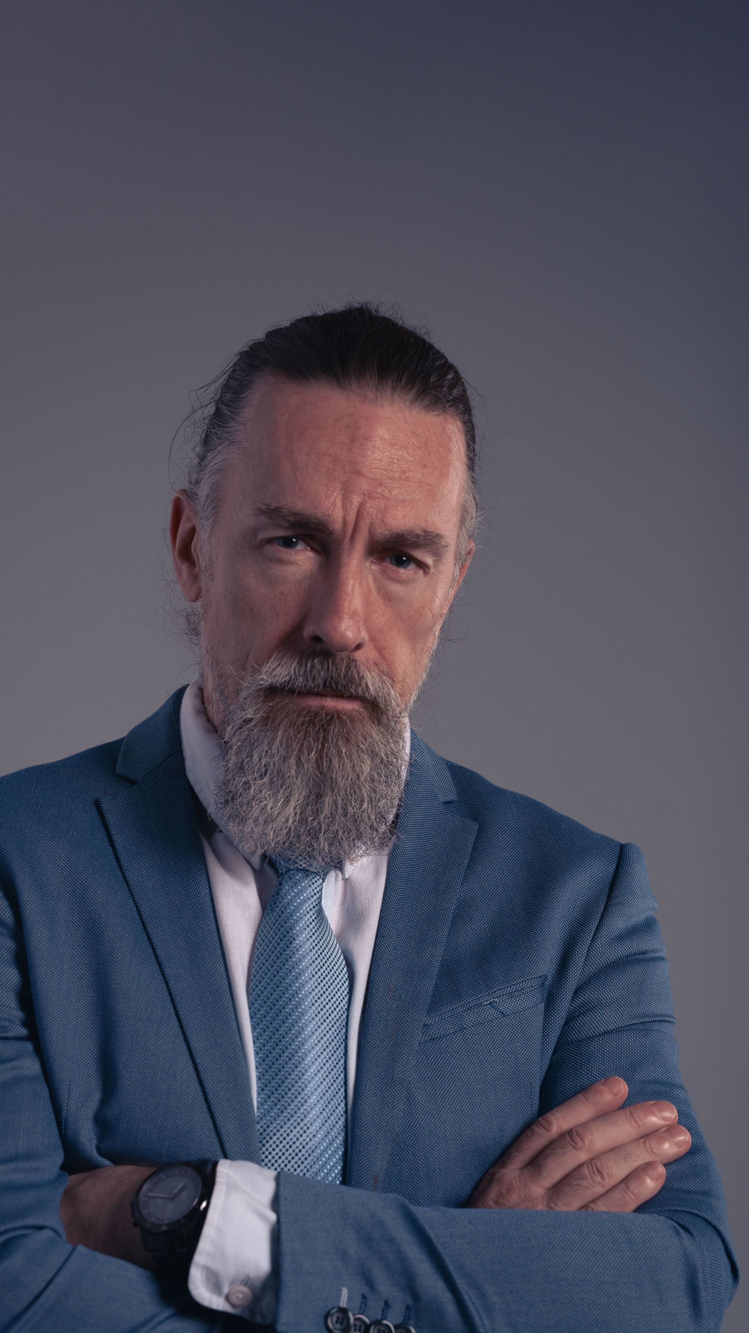 A distinguished middle-aged man with a gray beard in a blue suit and tie stands with arms crossed against a gray background.