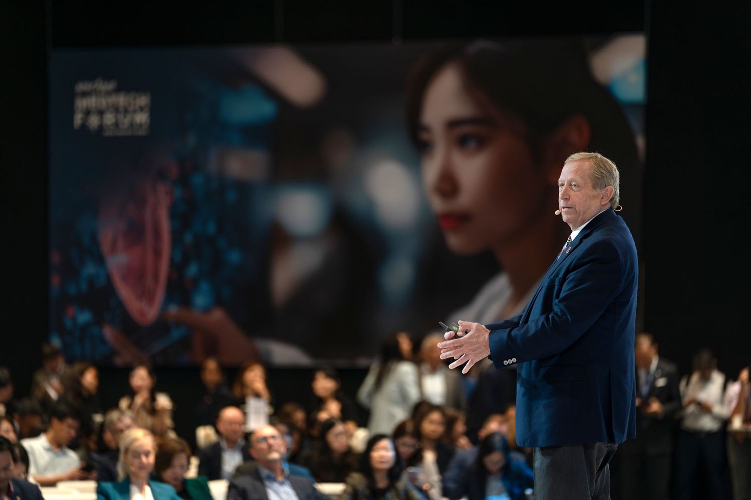 A speaker presents on stage at a forum event with a large screen displaying a womans face in the background.