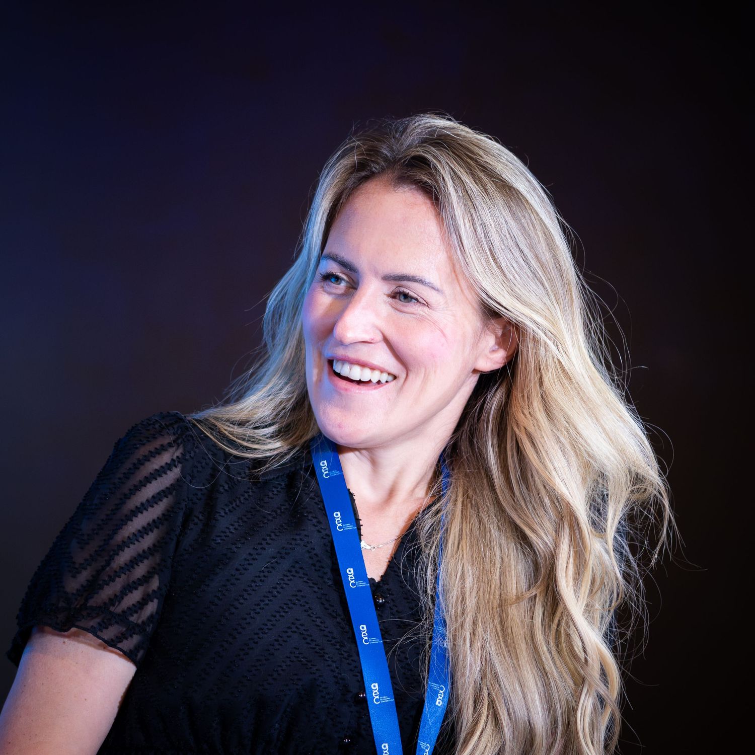 A smiling blonde woman wearing a black dress and blue lanyard at a professional event.