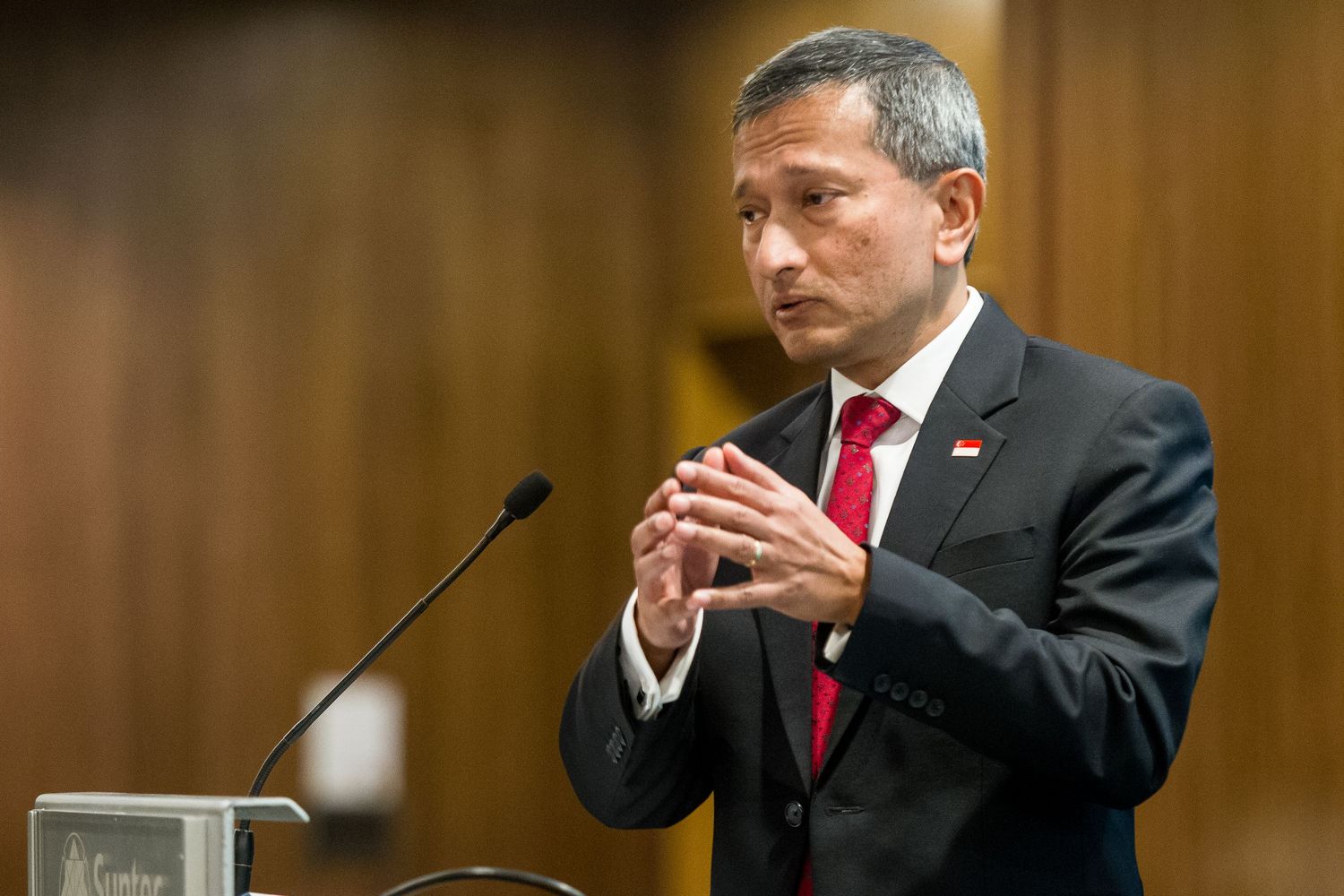Man in dark suit with red tie speaks at podium, gesturing with hands clasped in formal setting.