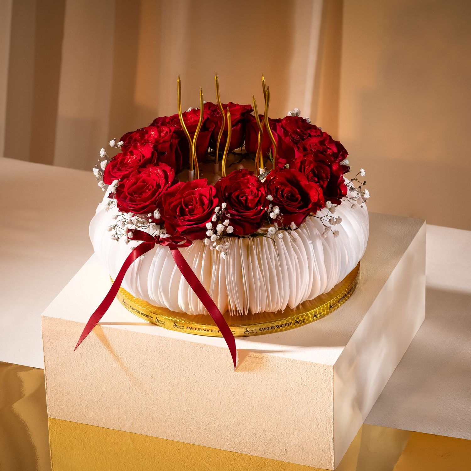Elegant floral cake topped with red roses, baby's breath, gold accents, and a red ribbon on a gold base.
