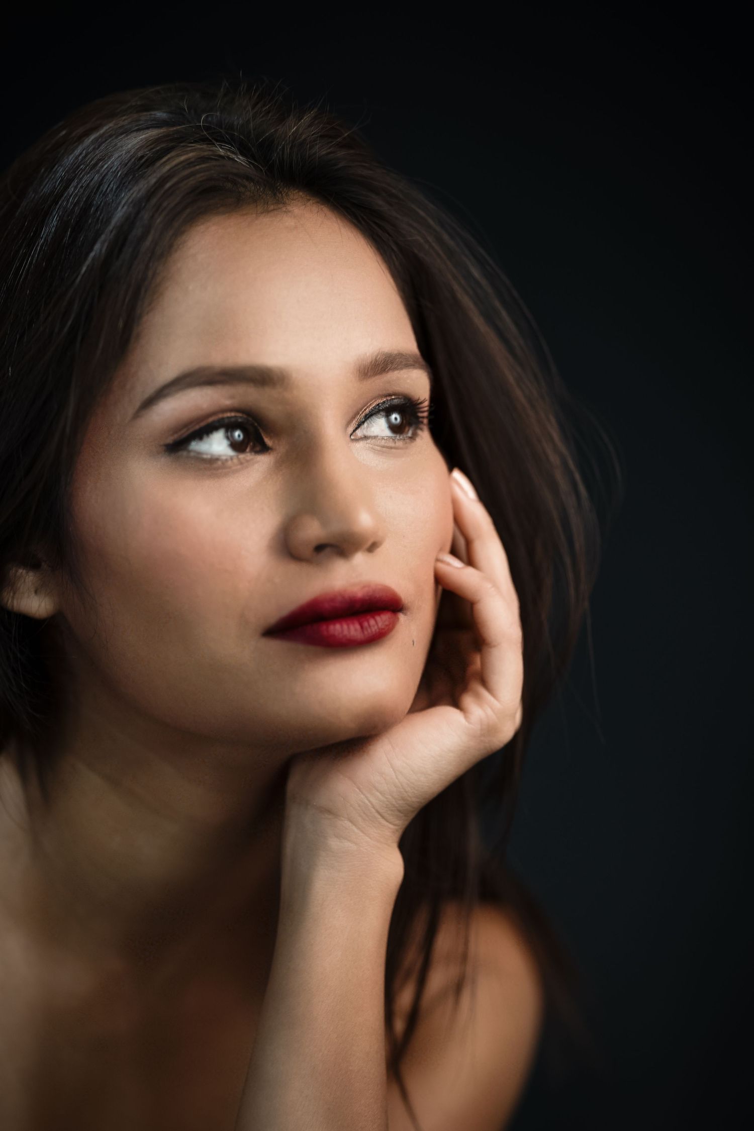 Young woman with dark hair and red lips resting chin on hand, gazing upward against dark background.