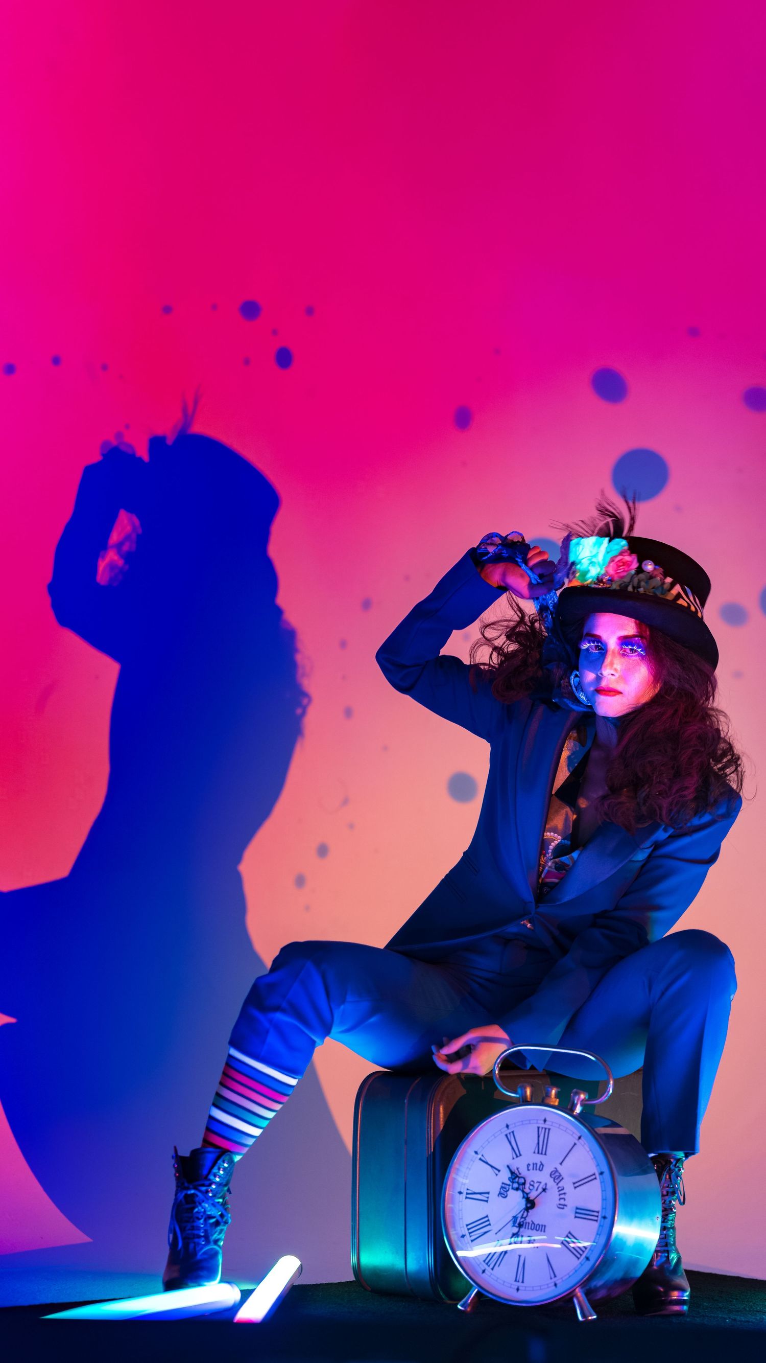 A woman in a Mad Hatter costume sits on a suitcase with a clock, posed against a vibrant pink and purple neon background.