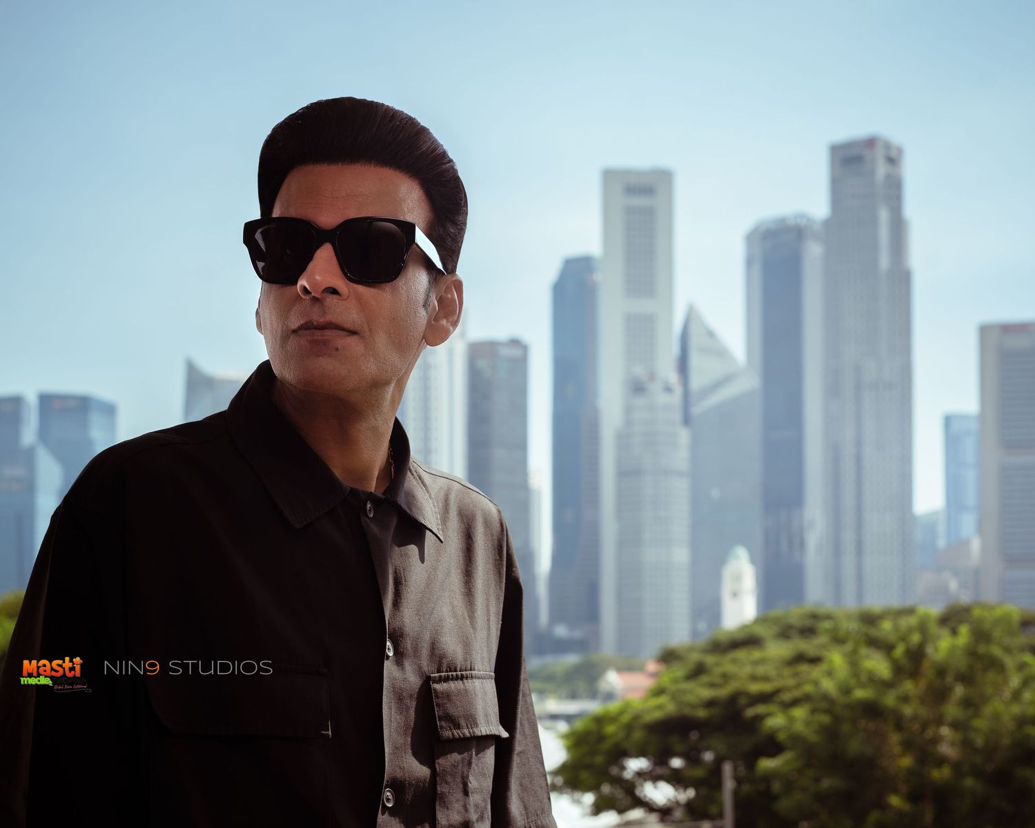 Man in sunglasses and dark jacket poses against Singapore city skyline on a clear day.