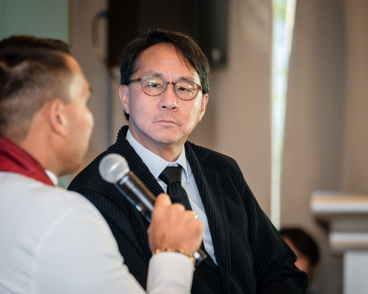 Two men in formal attire engage in conversation, one holding a microphone at an indoor event.