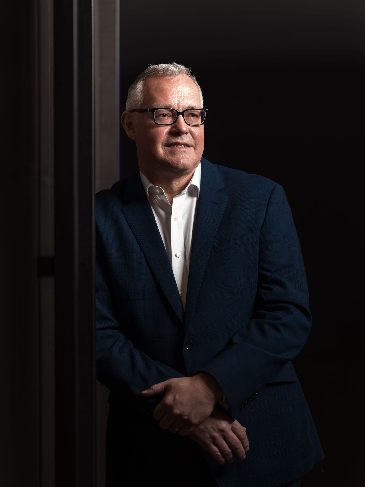 Middle-aged man with glasses in navy blazer posing confidently against dark background in professional portrait.