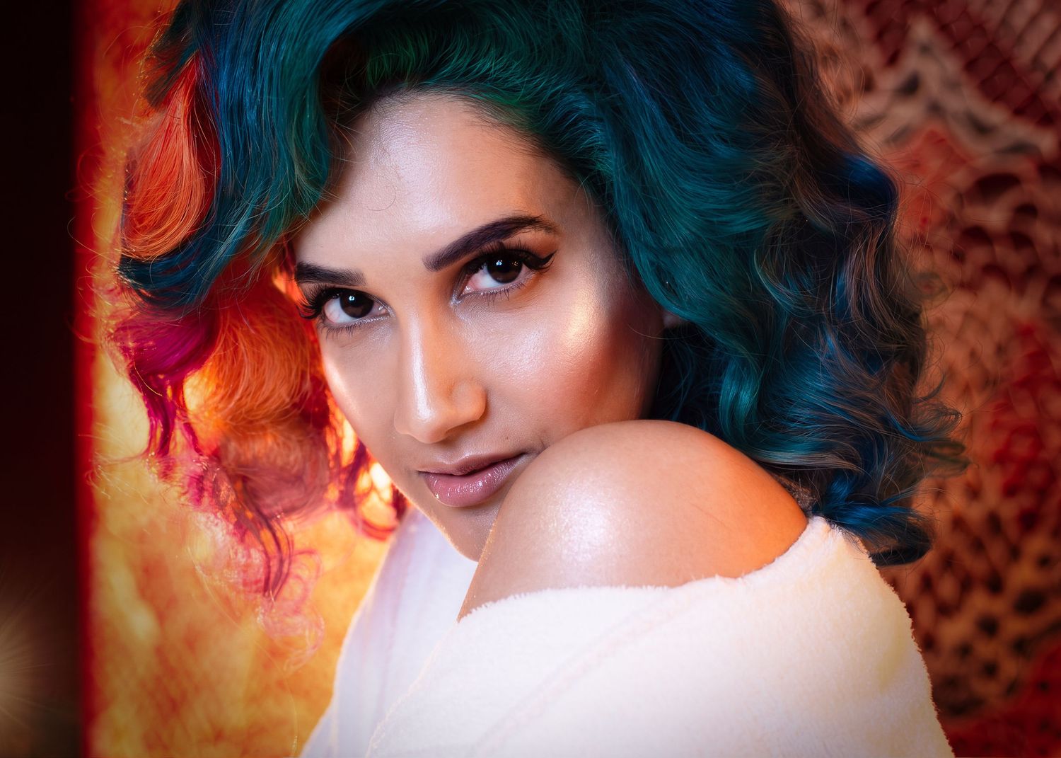 Woman with vibrant teal, orange, and pink curly hair poses in a white off-shoulder top against a warm background.