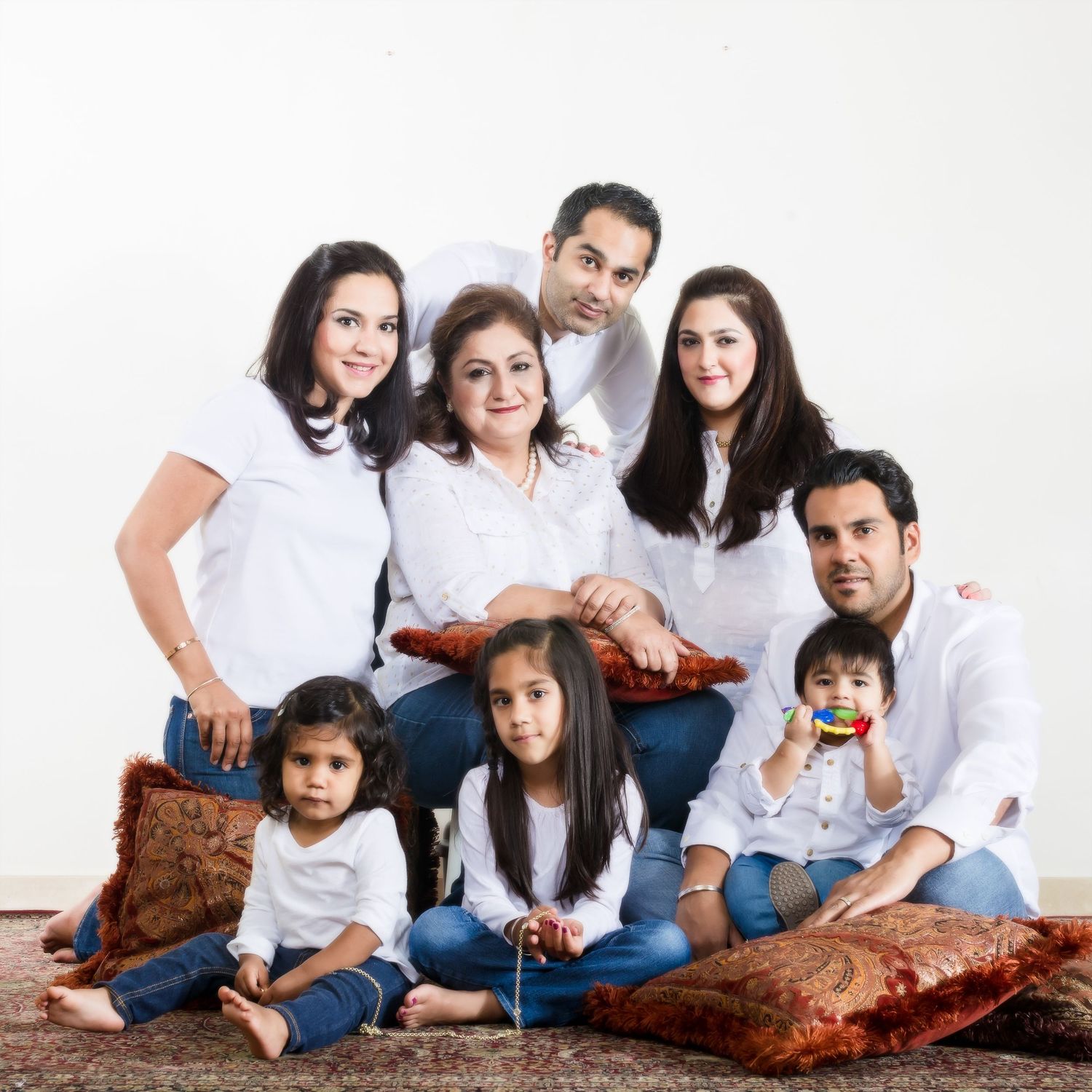 A multigenerational South Asian family of seven poses together in white outfits on a decorative rug for a studio portrait.