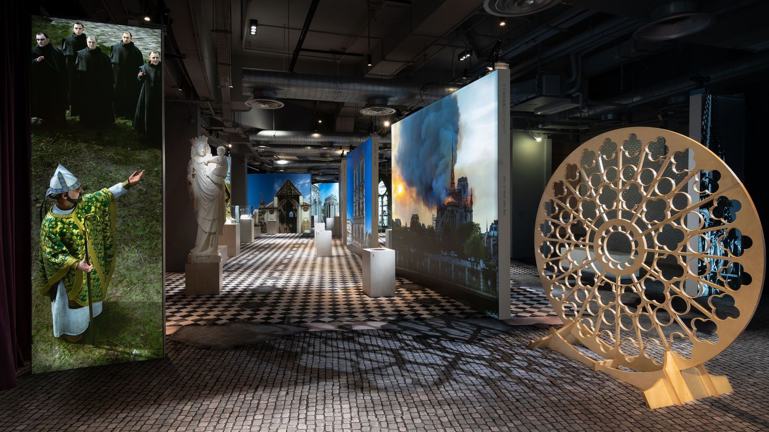 Immersive art installation with large-scale projections, sculptural figures, and a decorative wooden wheel in a dimly lit gallery.