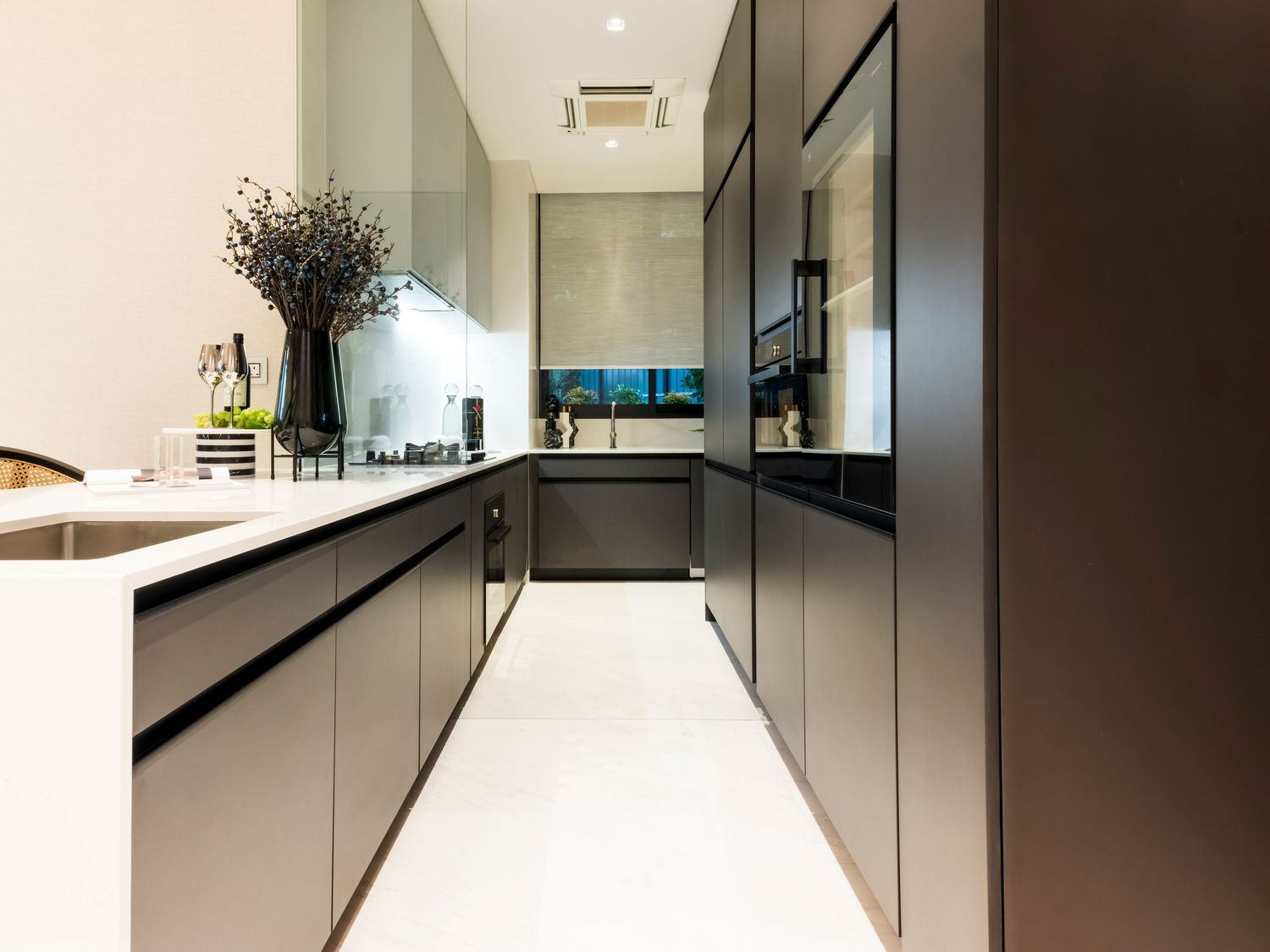 Modern galley kitchen with dark cabinetry, white countertops, and large format white floor tiles in a sleek minimalist design.