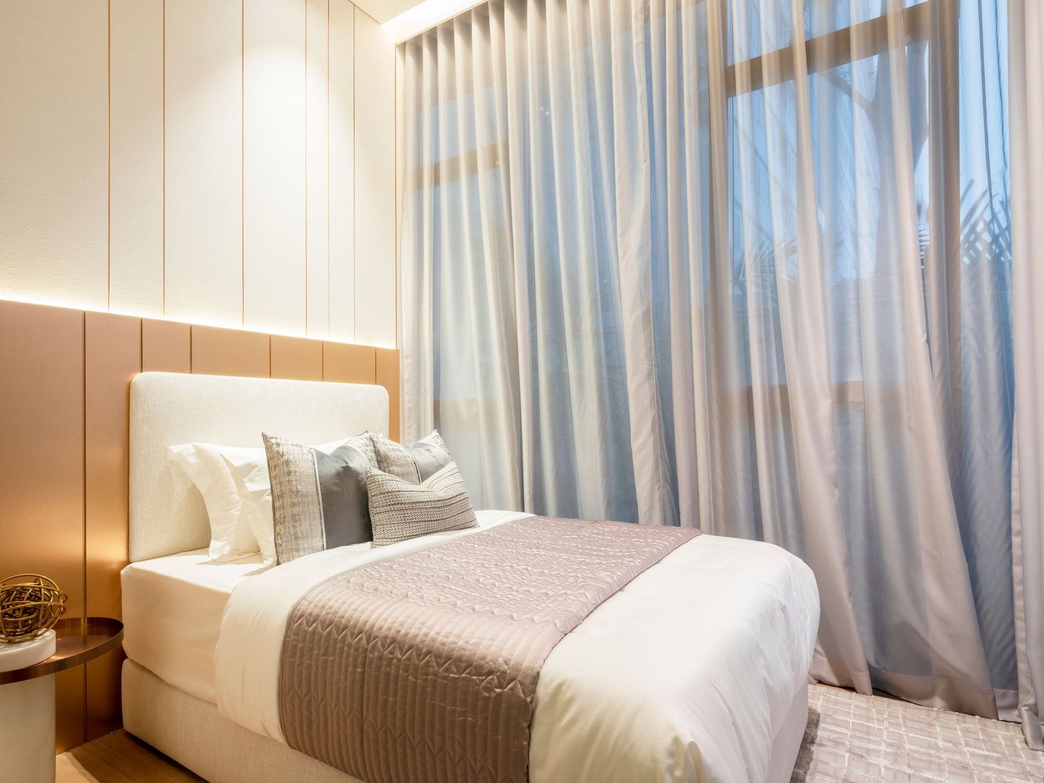 Elegant bedroom with upholstered headboard, decorative pillows, neutral tones, and sheer curtains by a sunlit window.