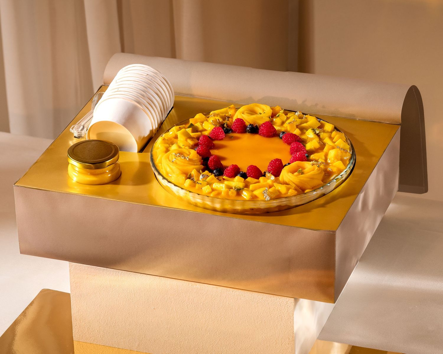 Golden tart topped with fresh raspberries and yellow flowers on a gold serving board with stacked white plates.