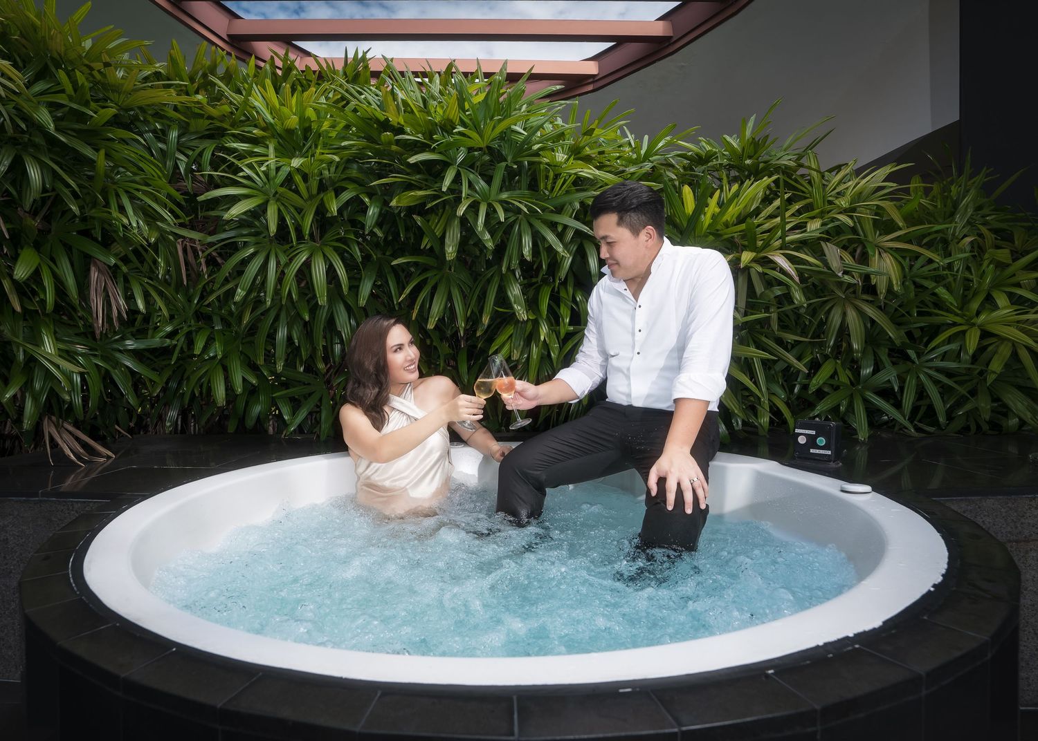 Couple relaxing in an outdoor hot tub jacuzzi surrounded by lush green tropical plants, sharing drinks together.