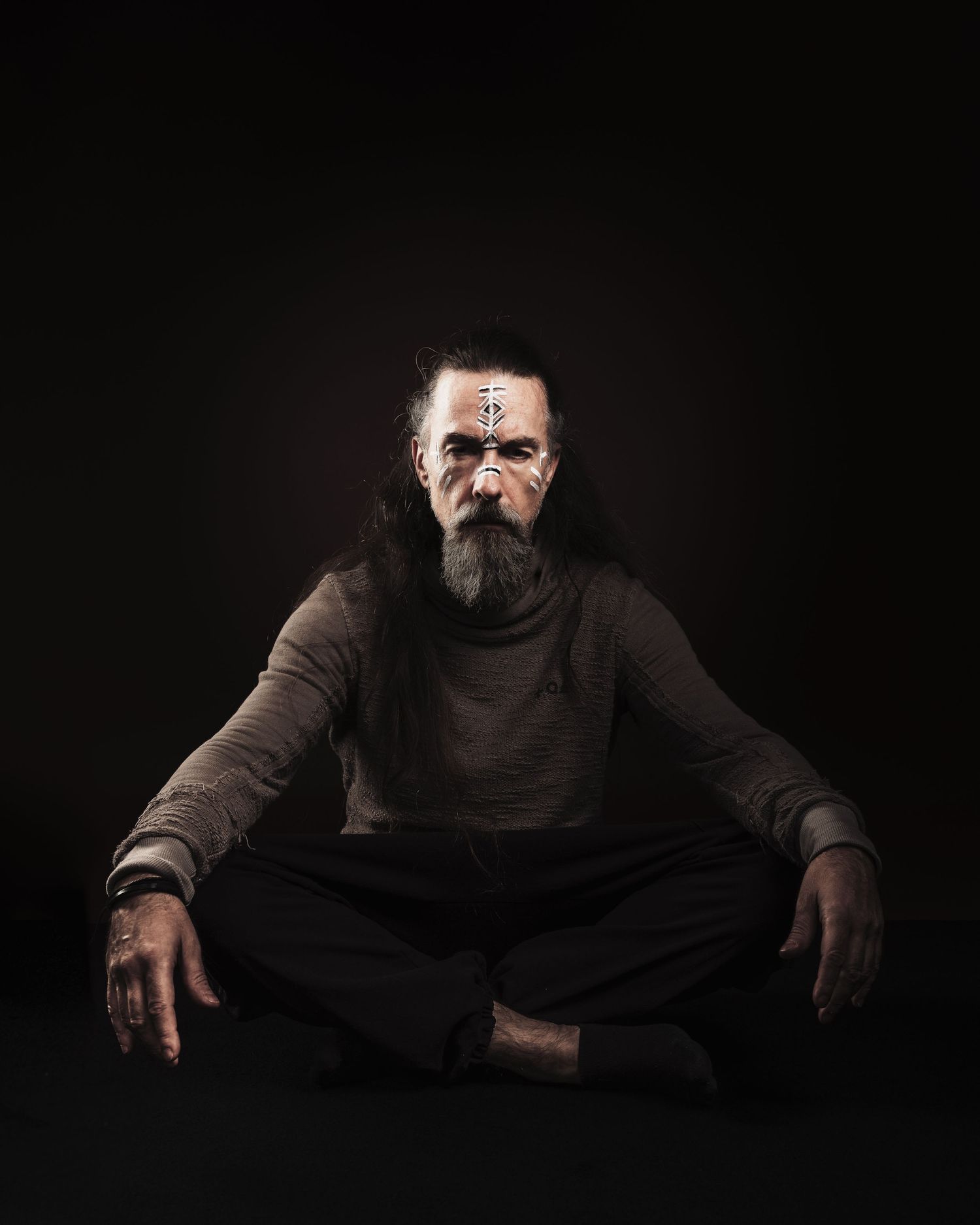 Bearded man with long hair and face paint sits cross-legged in meditation pose against dark background.