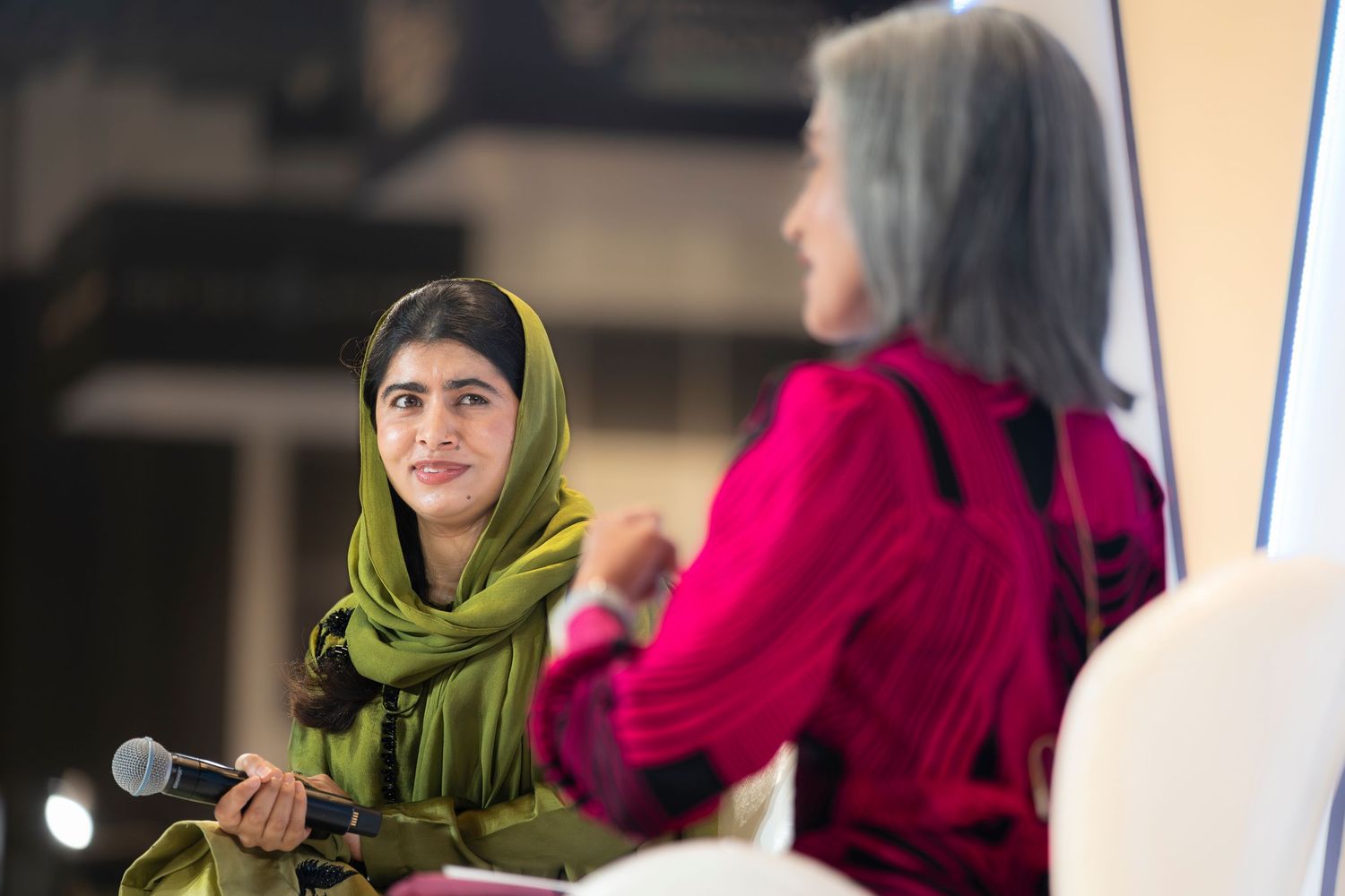 Woman in green hijab holding microphone speaks at panel discussion event with another woman in pink jacket.