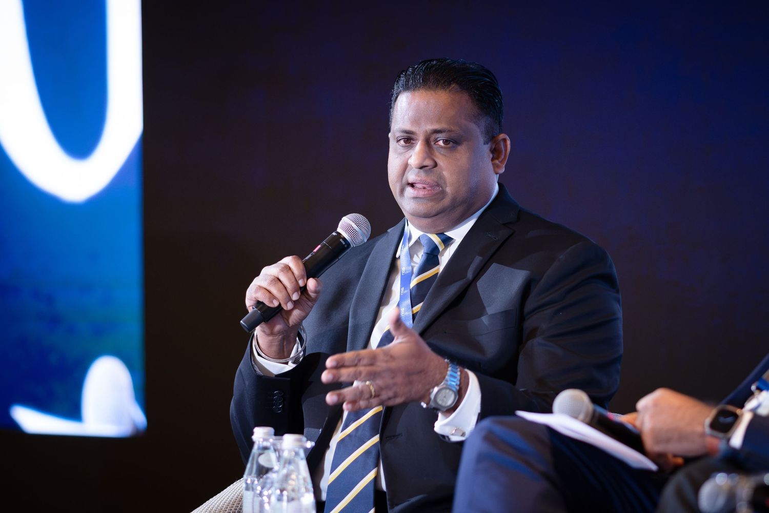 A man in a suit speaking at a panel discussion, holding a microphone against a dark blue background.