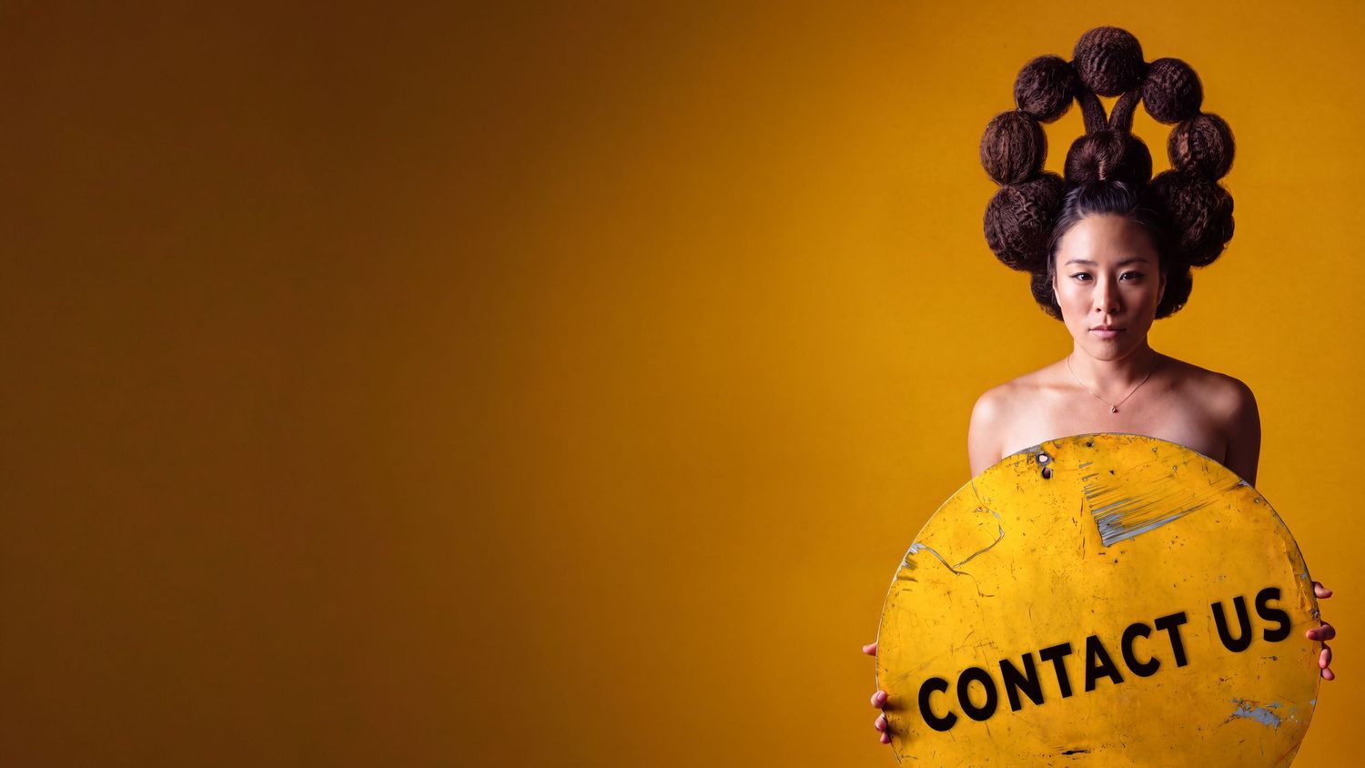 Woman with elaborate updo hairstyle holding a yellow 'Contact Us' sign against a golden background.