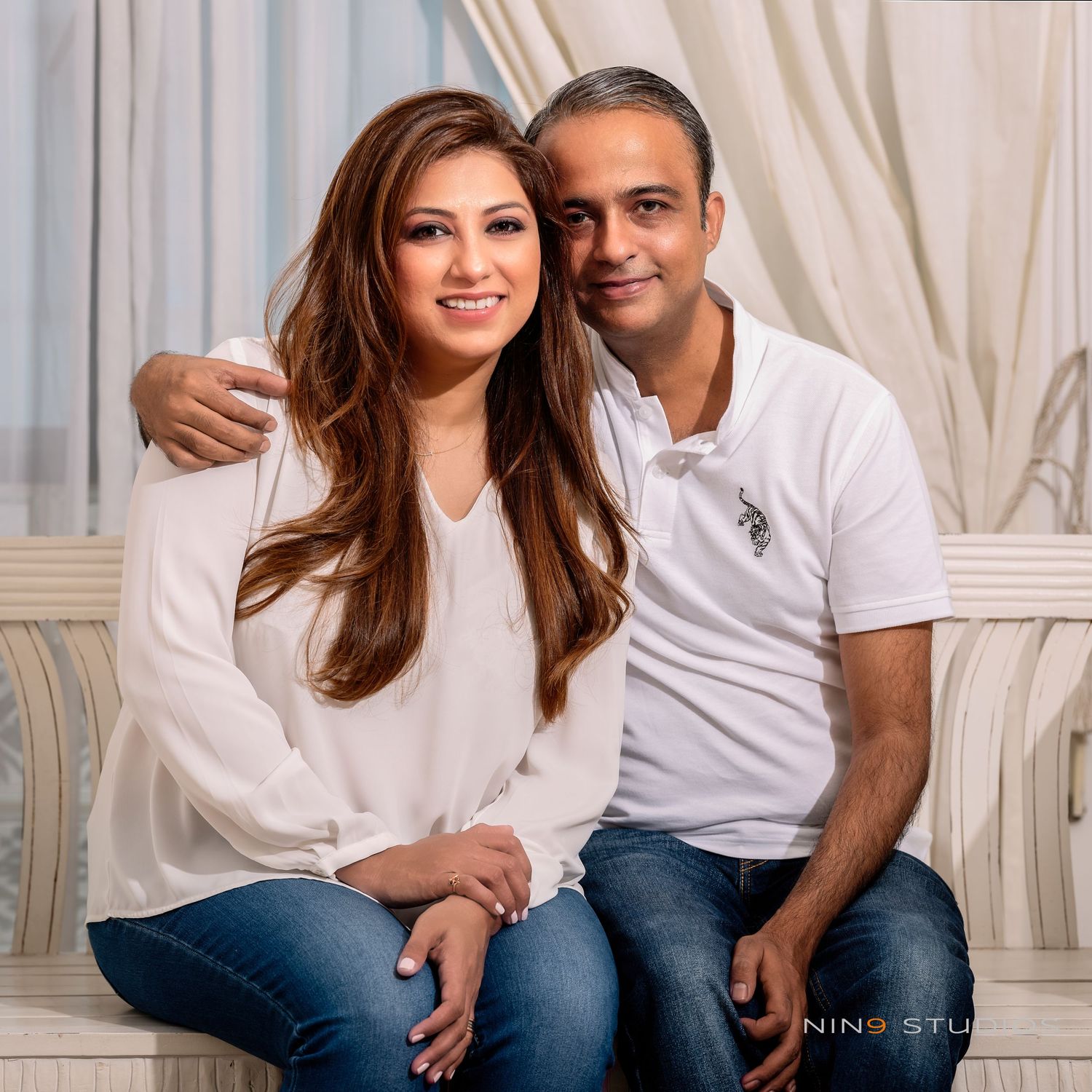 A smiling couple in white tops and jeans pose together on a couch in a bright, elegant studio setting.