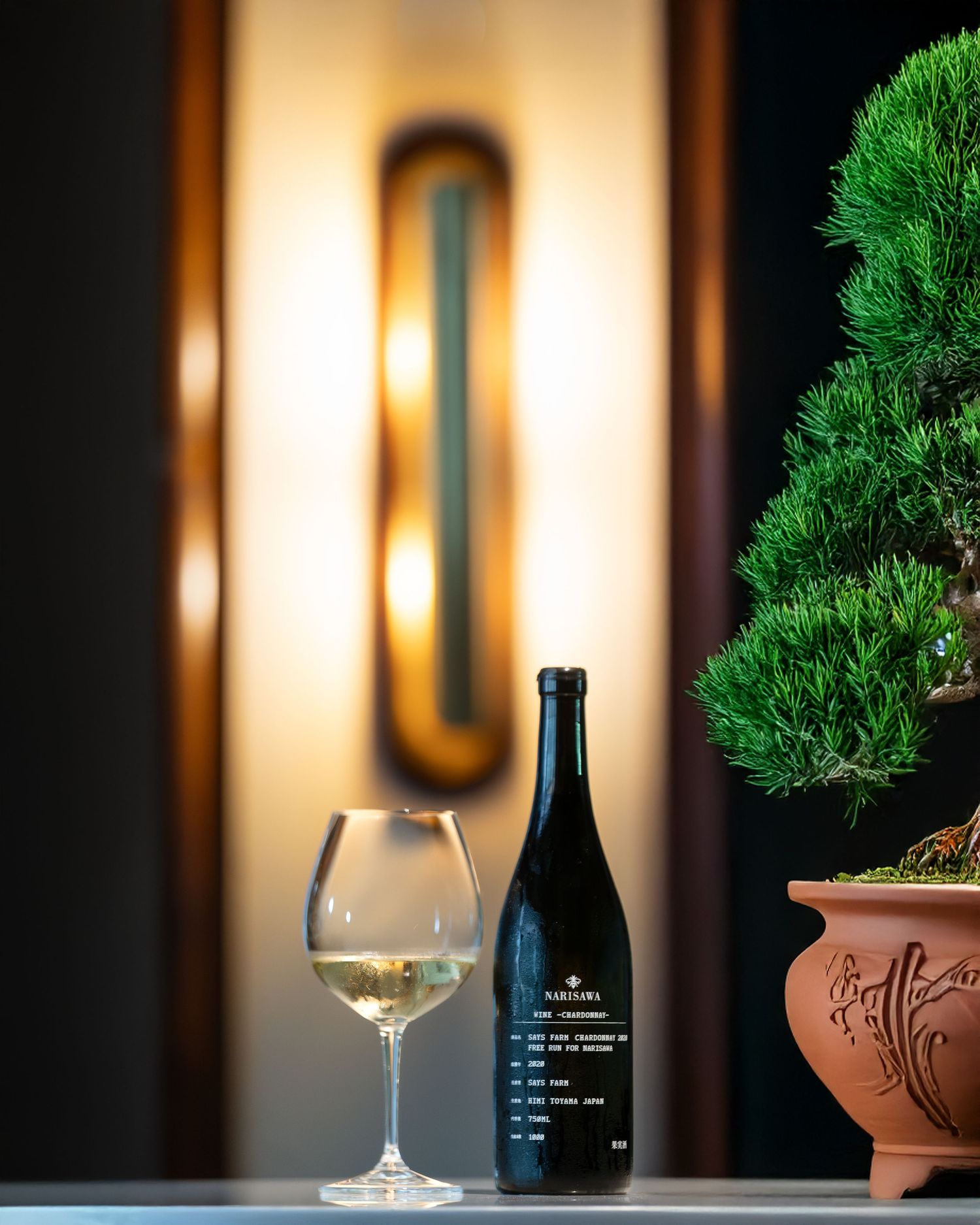Elegant white wine bottle beside a glass of white wine and bonsai tree, with warm wall sconce lighting.