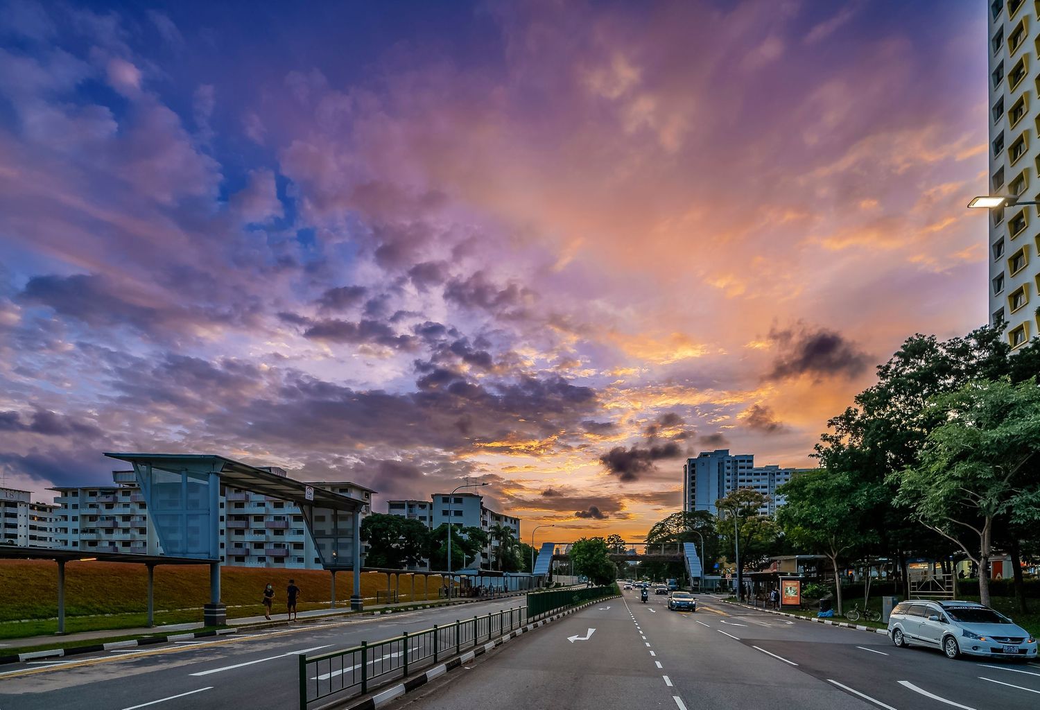 Urban road at sunset with dramatic purple and orange clouds over city buildings and lush trees lining the street.