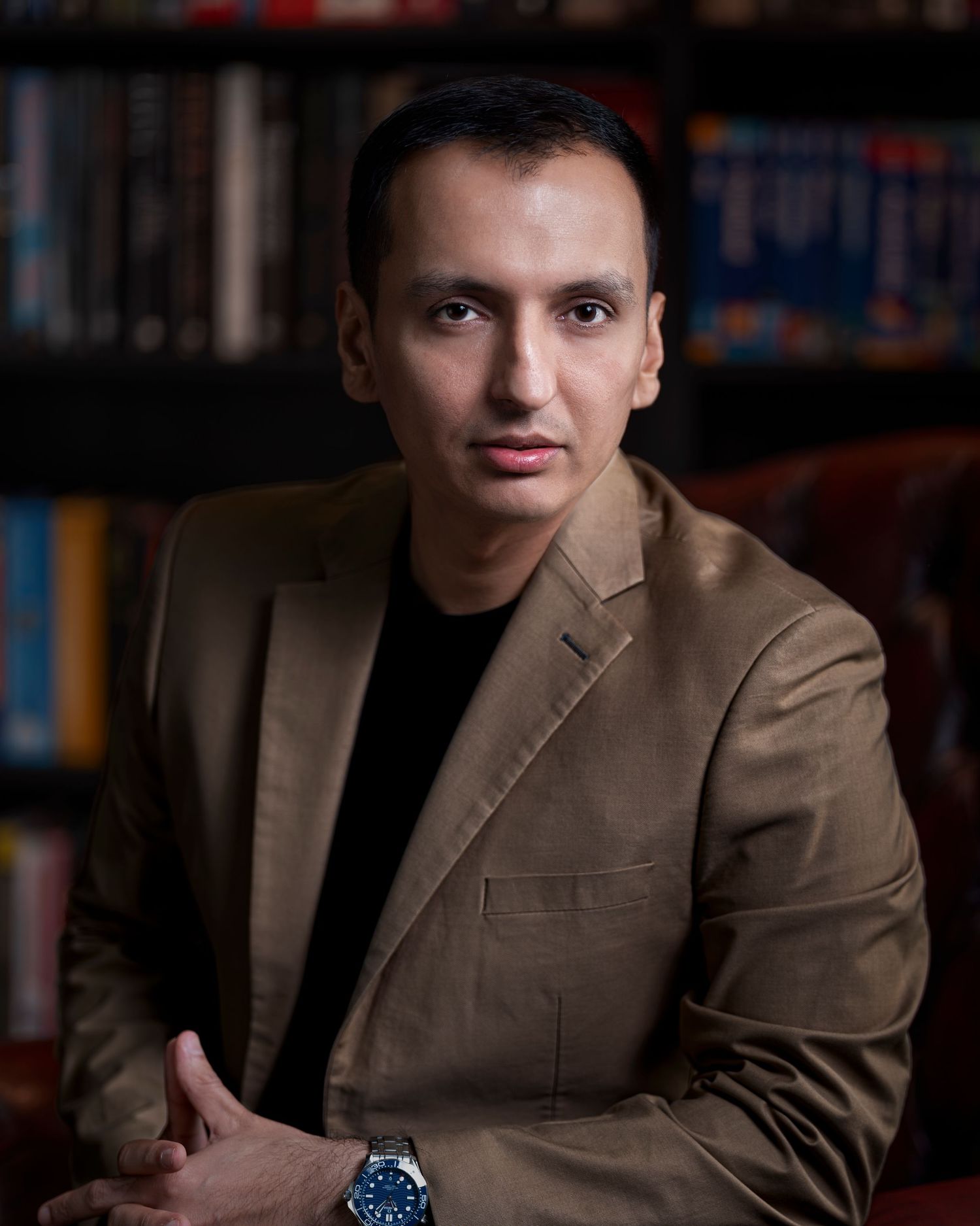 Professional man in tan blazer sits confidently before bookshelf, wearing a watch, in a formal portrait photo.