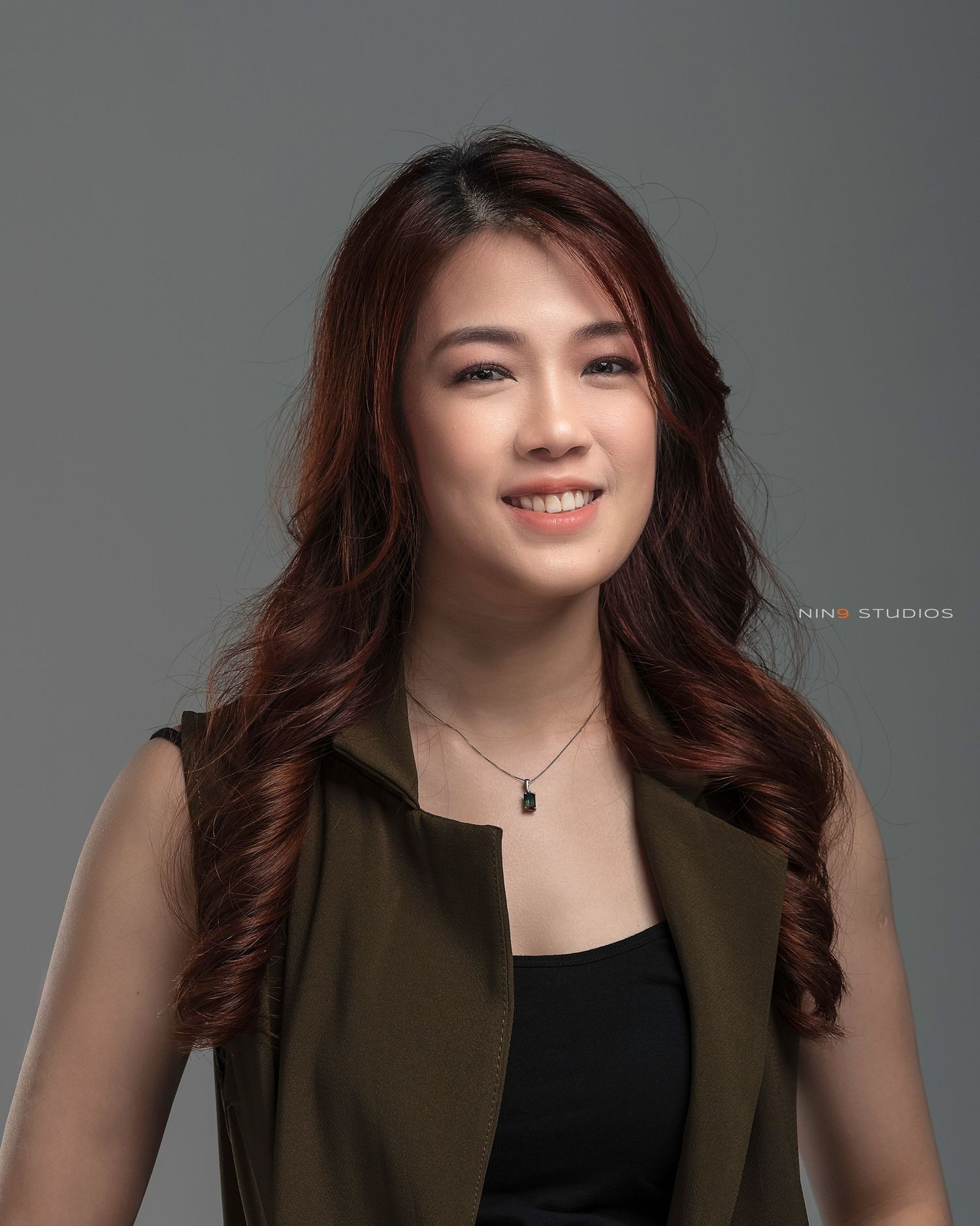 Professional headshot of a young Asian woman with long wavy auburn hair, wearing an olive vest over a black top.