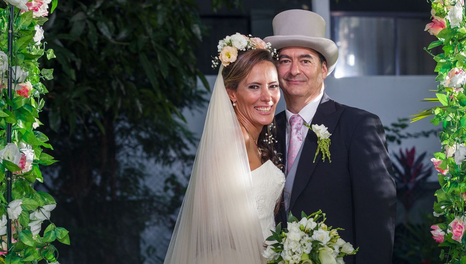 A bride and groom pose together under a floral arch, she in a white gown with veil, he in a gray top hat.