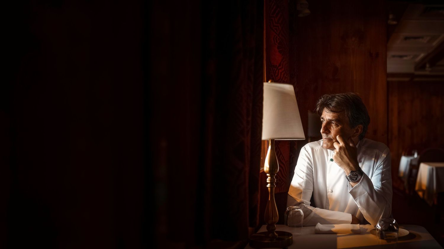Thoughtful man sitting alone at dimly lit restaurant table beside a warm glowing lamp in dark ambiance.