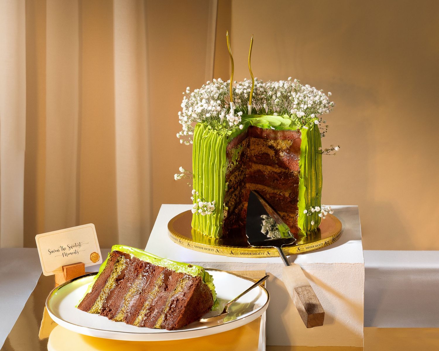 Elegant layered cake with green frosting and white flowers, sliced to reveal chocolate layers, served on a golden plate.
