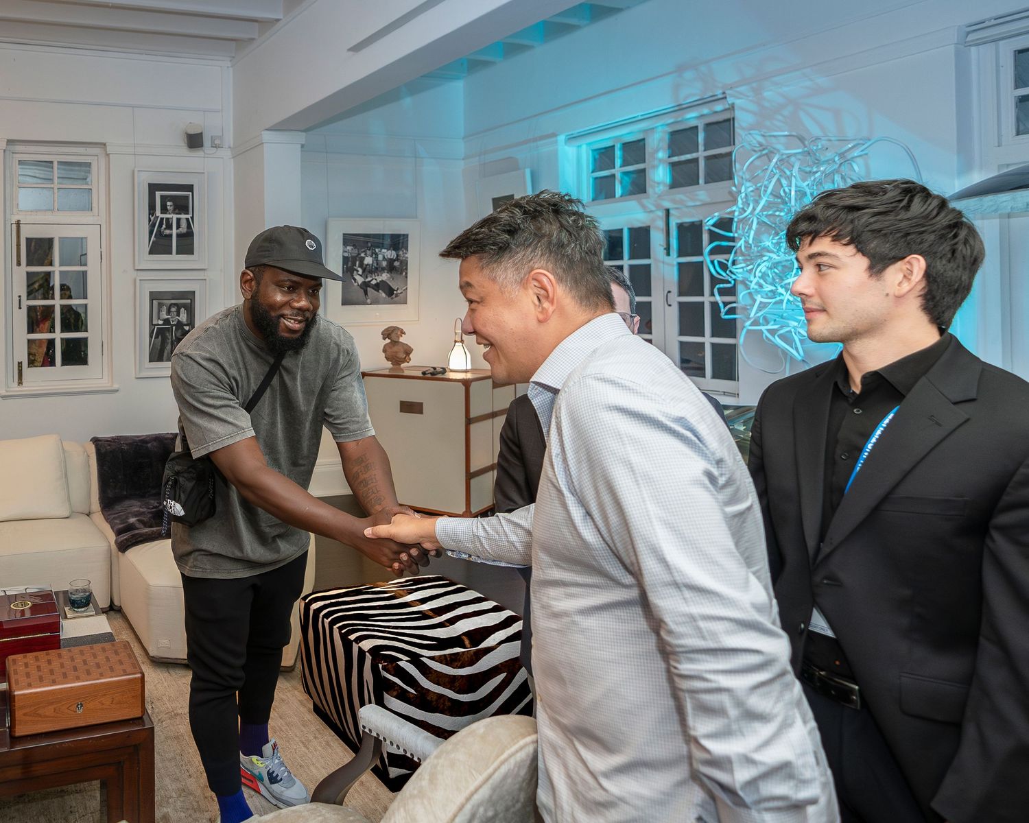 Two men shake hands warmly in an elegant room while a third man stands nearby observing.