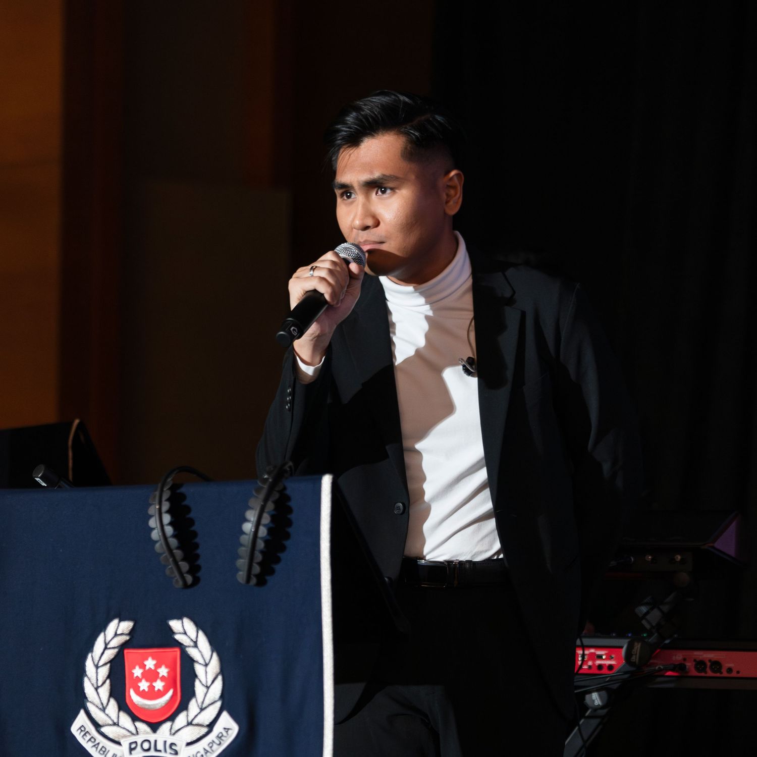 Young man in black blazer singing into microphone at Singapore Police Force event on stage.