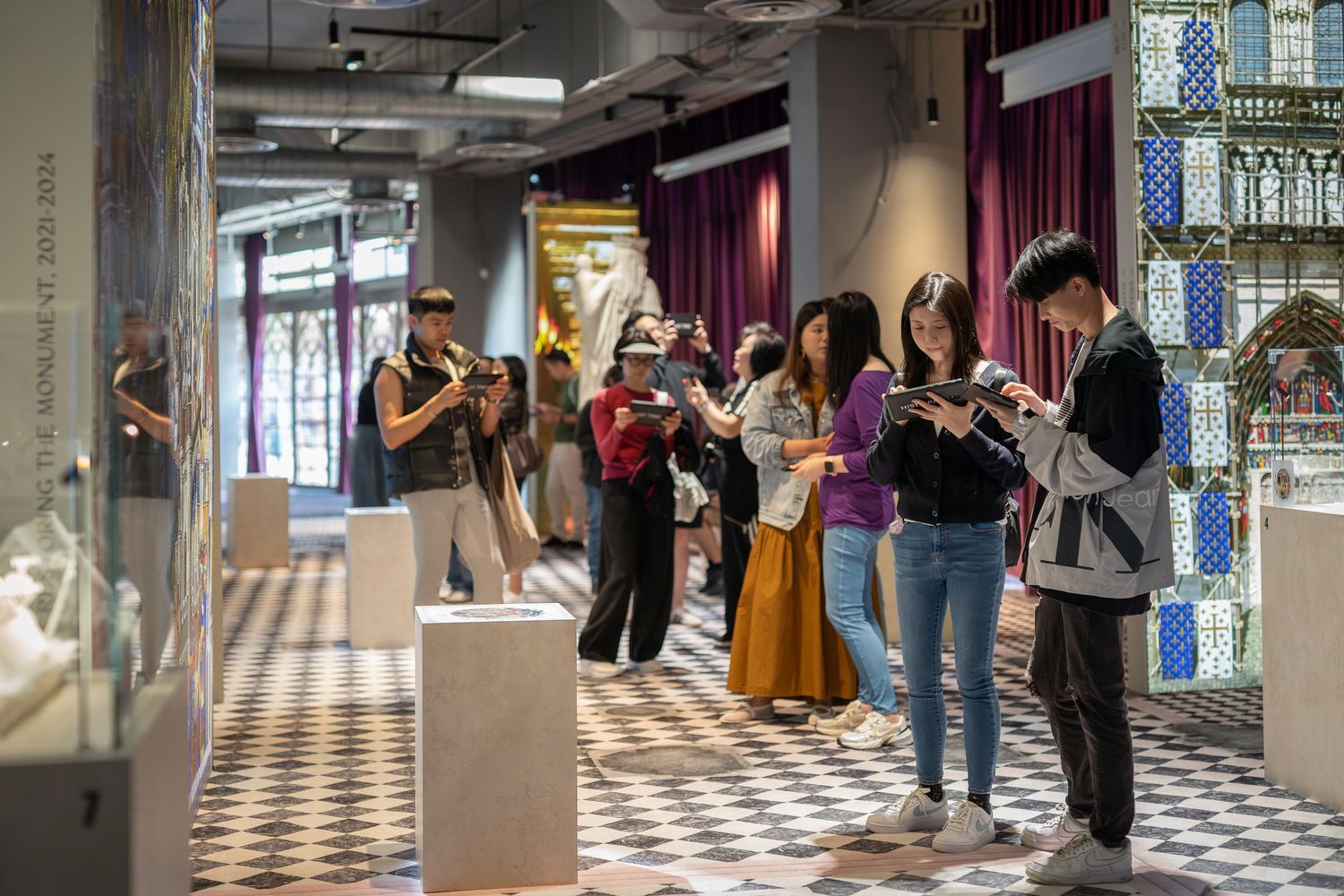 Museum visitors browse artifacts and sculptures in gallery with decorative tiled flooring.
