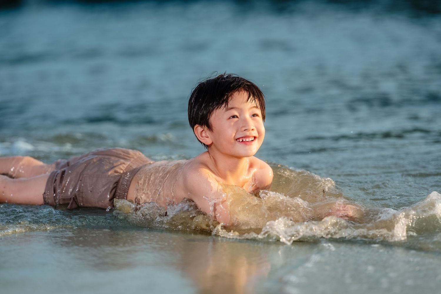 A smiling Asian boy lying in shallow ocean waves on a sandy beach, enjoying a sunny day at the sea.