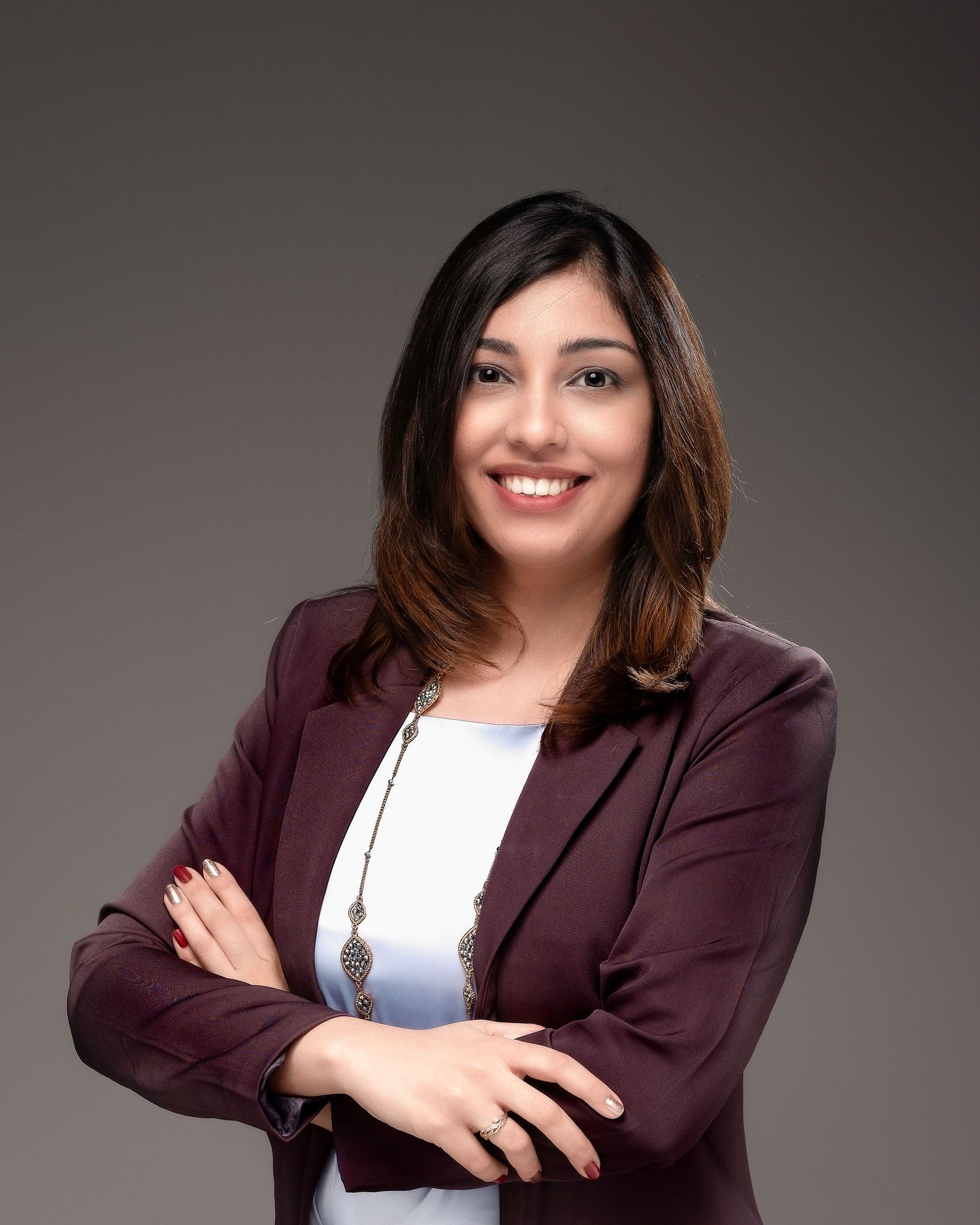 Professional woman in burgundy blazer smiling with arms crossed against gray background.