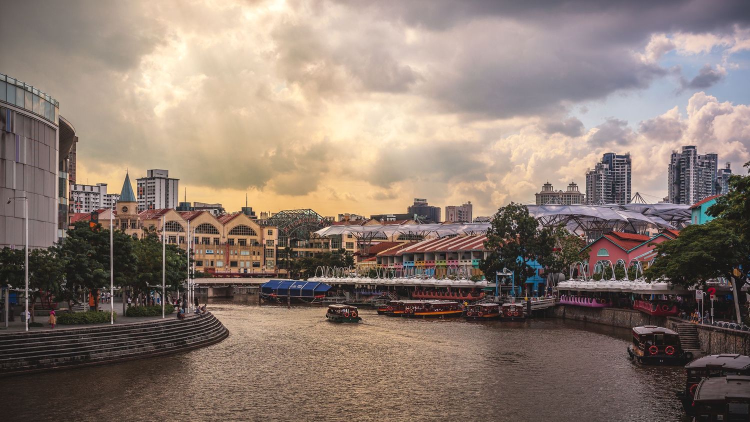 Scenic Singapore River with colorful Clarke Quay buildings, boats, and dramatic cloudy sky at sunset.