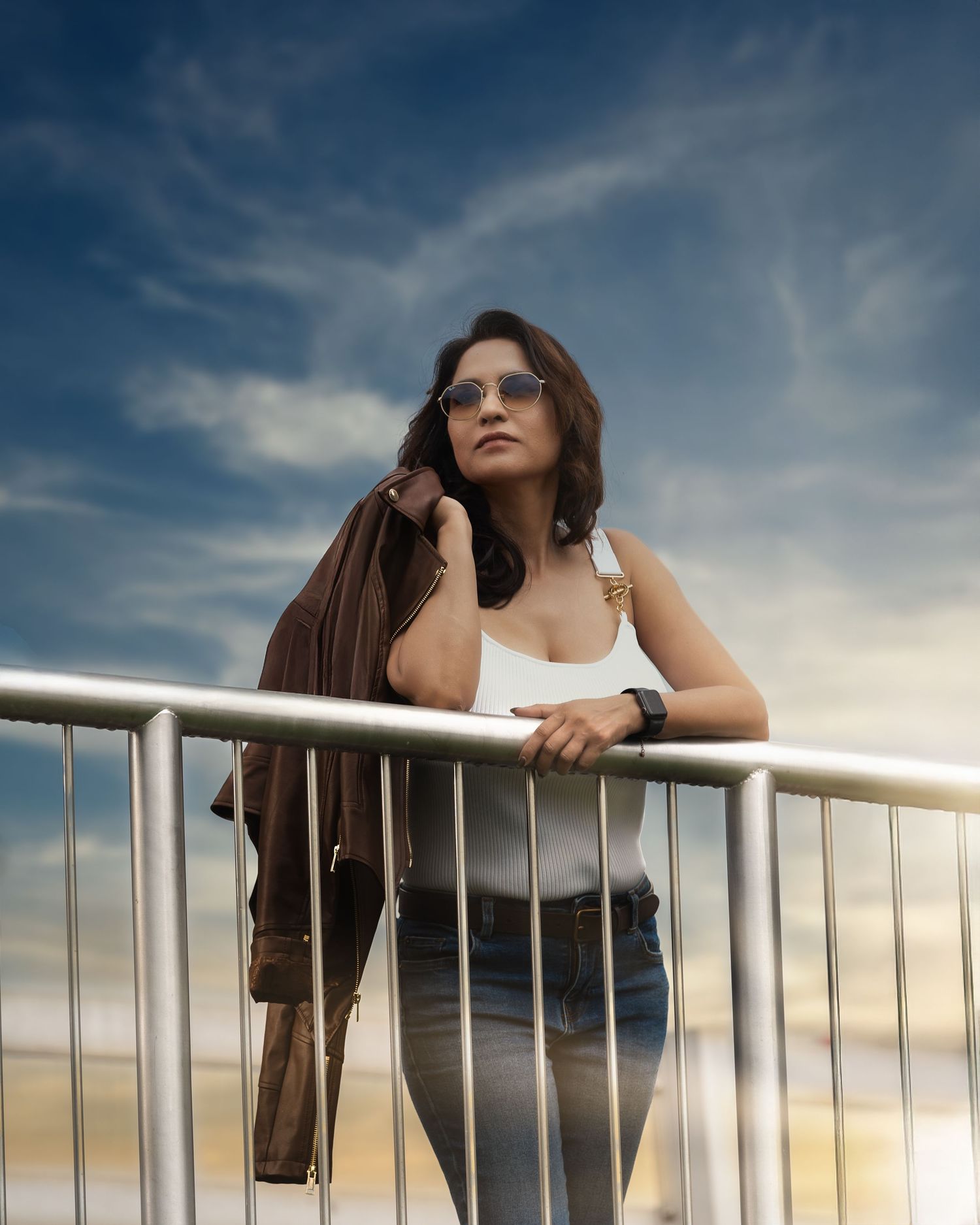 Woman in sunglasses leaning on metal railing, holding brown jacket, wearing white top and jeans under dramatic cloudy sky.