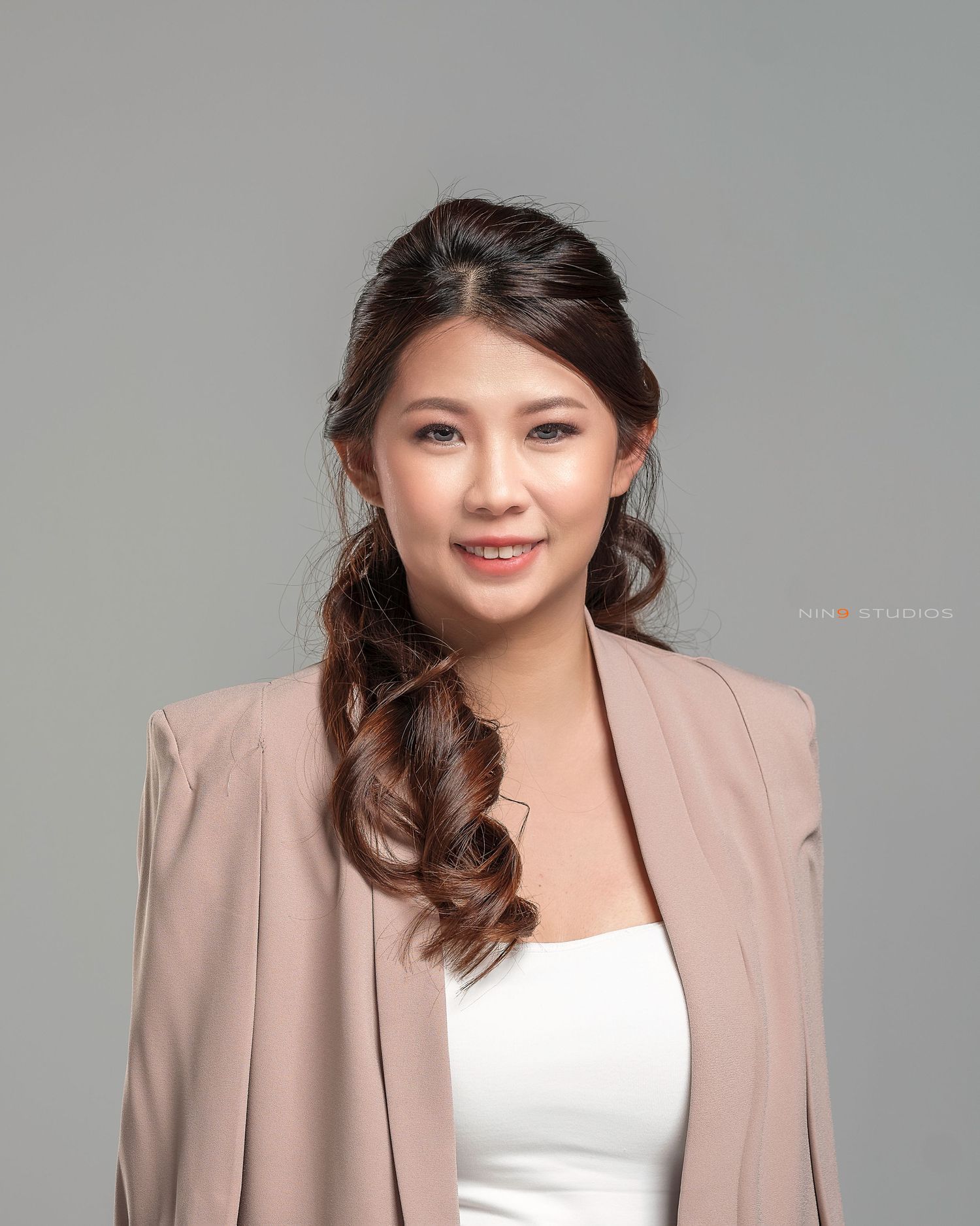 A young Asian woman in a blush blazer and white top with a side ponytail poses against a gray background.