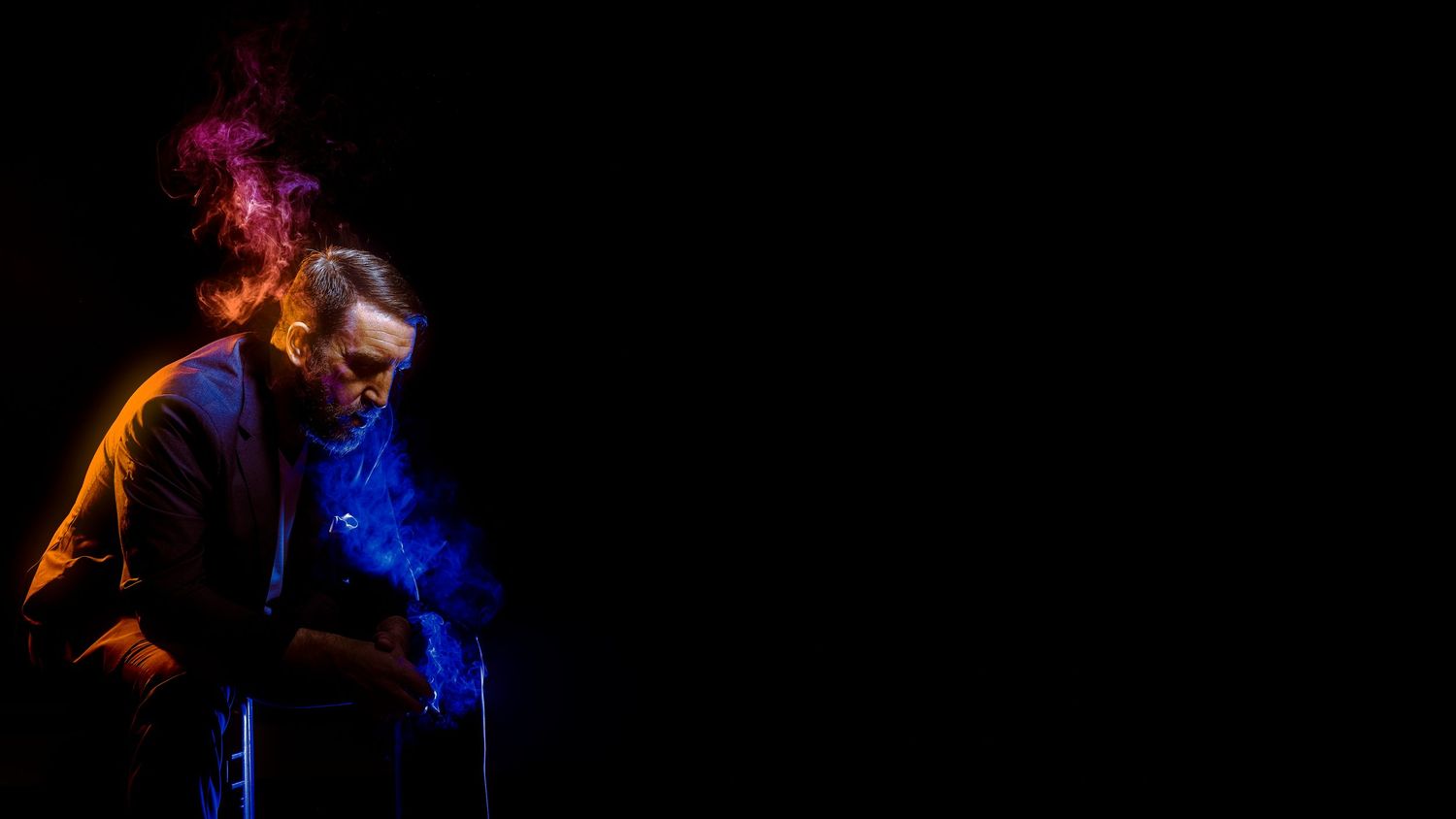 Artistic portrait of man surrounded by colorful smoke against black background in dark studio.