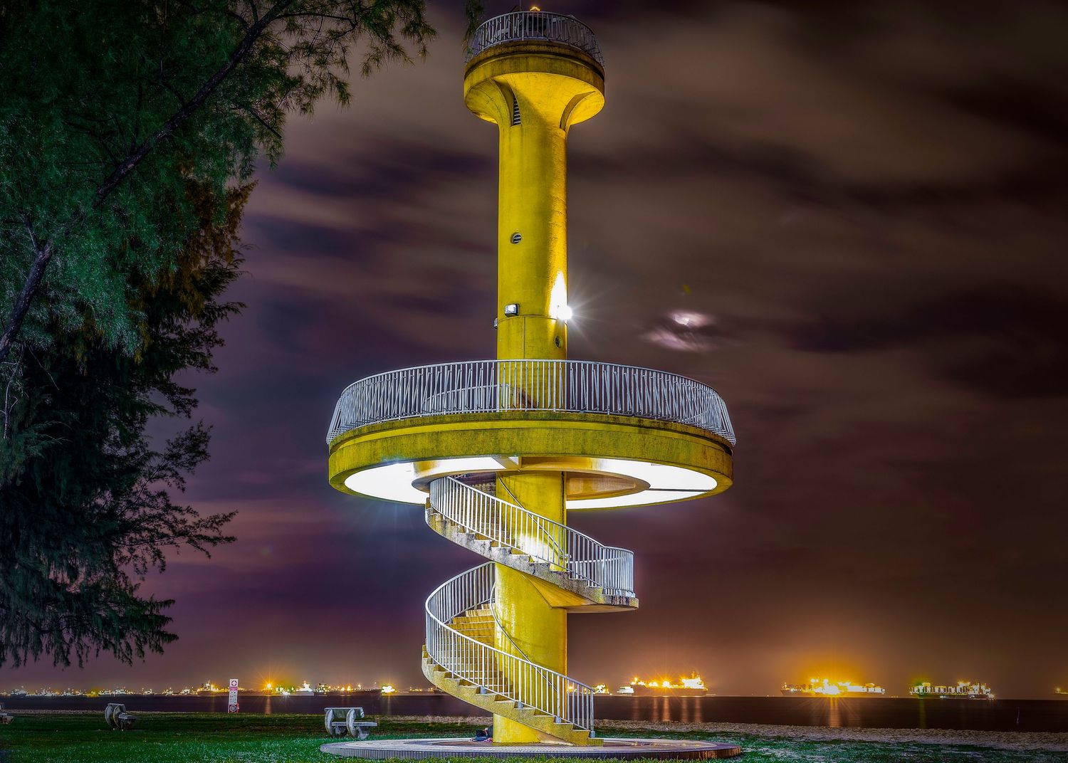 Yellow spiral staircase tower illuminated at night against a dramatic cloudy sky with city lights in the background.