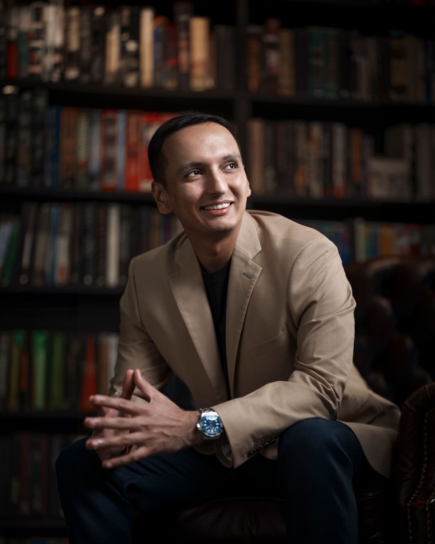 Smiling man in beige blazer sitting in front of bookshelf, looking up confidently with clasped hands.