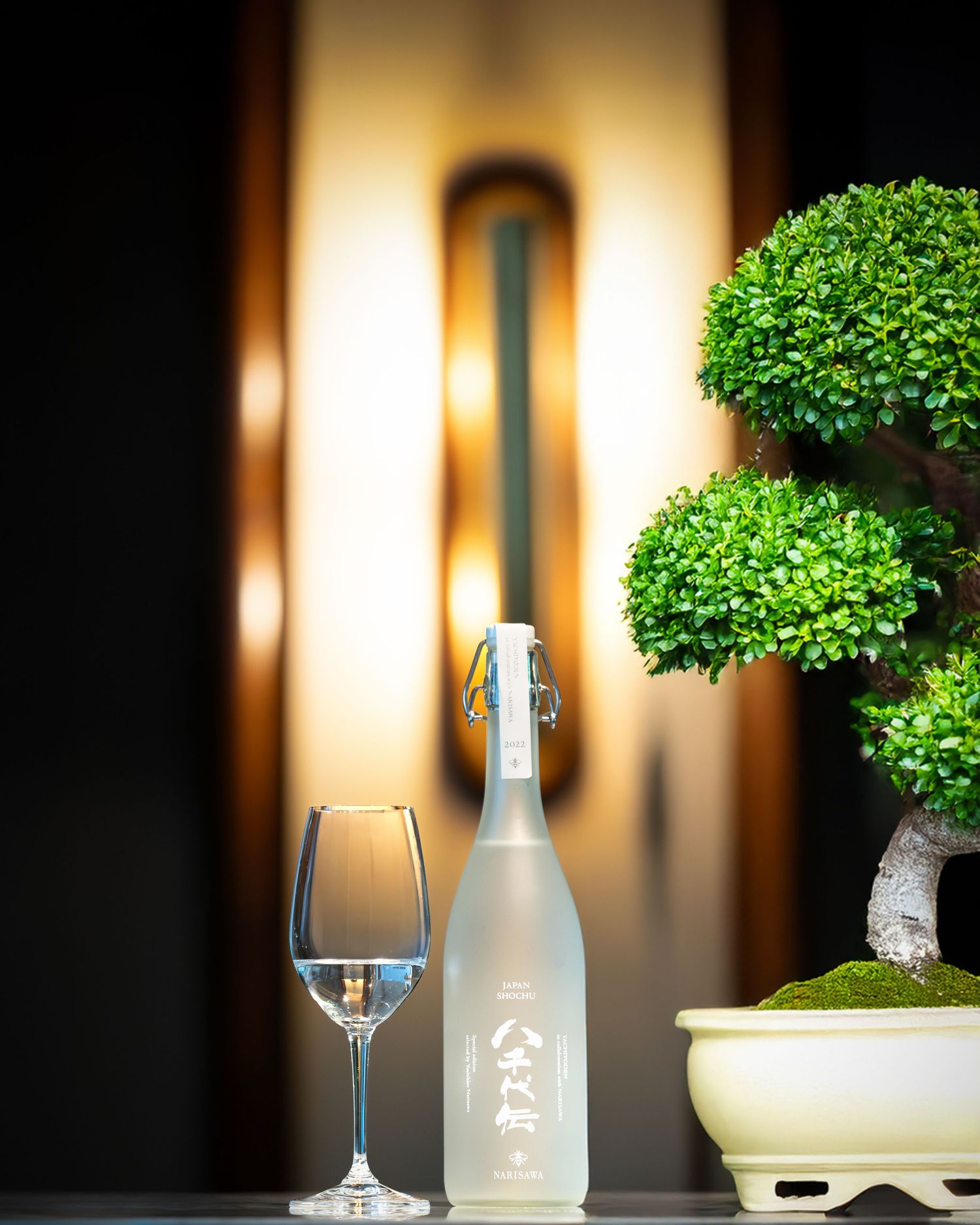 A frosted sake bottle beside a wine glass and bonsai tree, elegantly displayed against a warm glowing wall sconce.