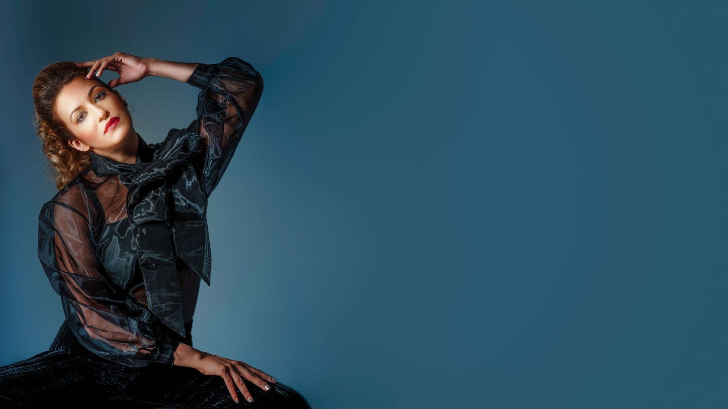 Woman in black sheer blouse posing elegantly against a teal blue background in a fashion portrait.