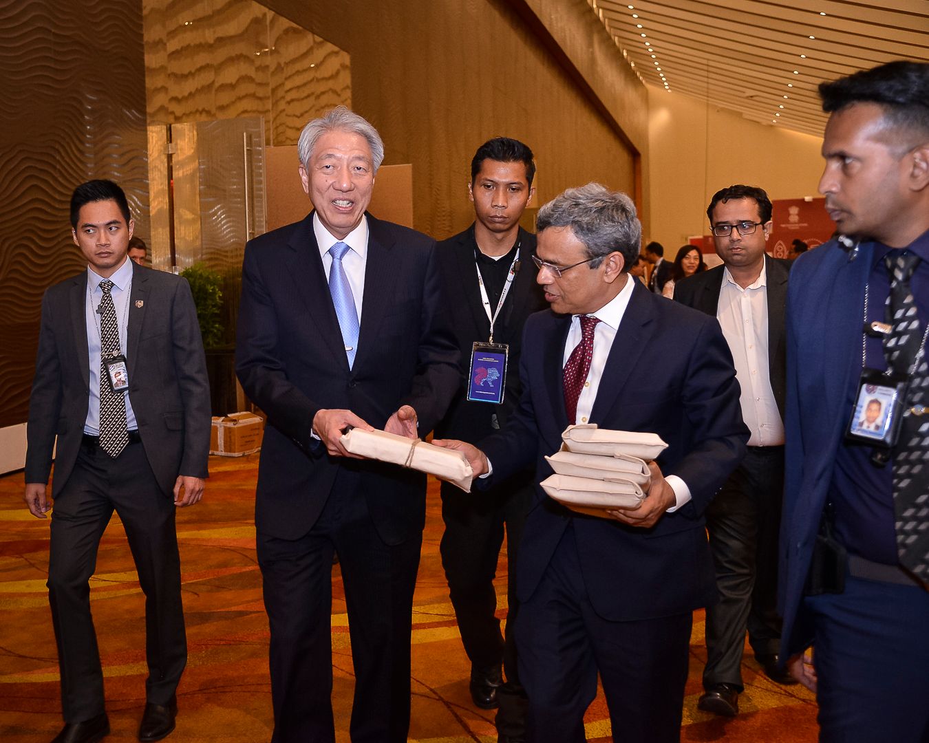Asian dignitaries exchange documents in an elegant convention center lobby surrounded by security personnel.